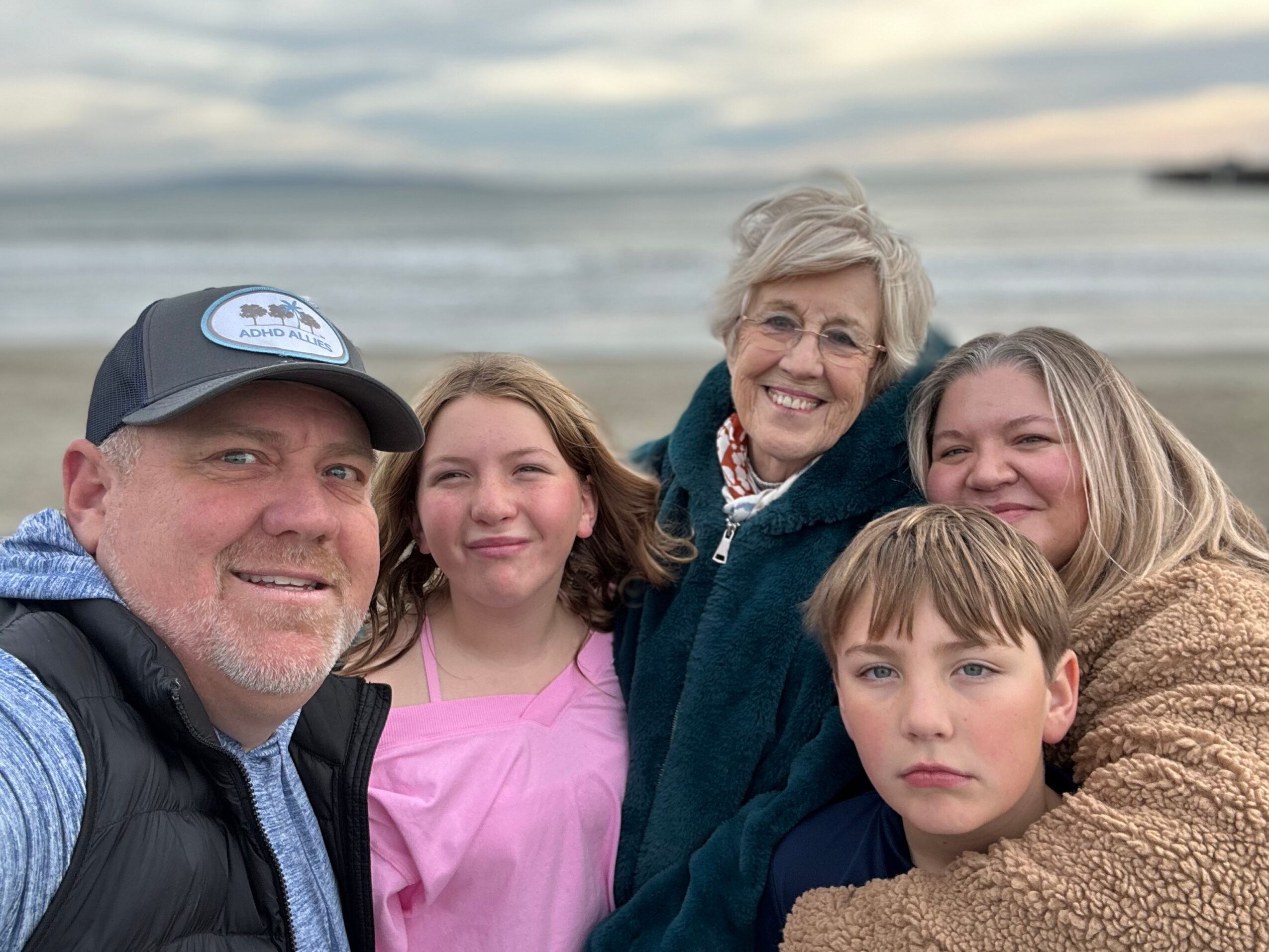 Group of five people smiling outdoors near water, including an older woman and children, with a cloudy sky background.