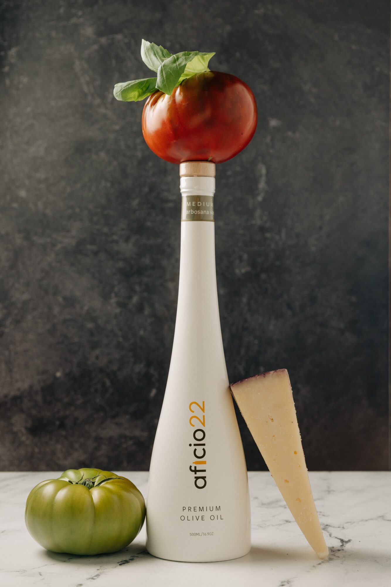 A bottle with a red apple on top, a green tomato, and a wedge of cheese on a marble surface against a dark background.