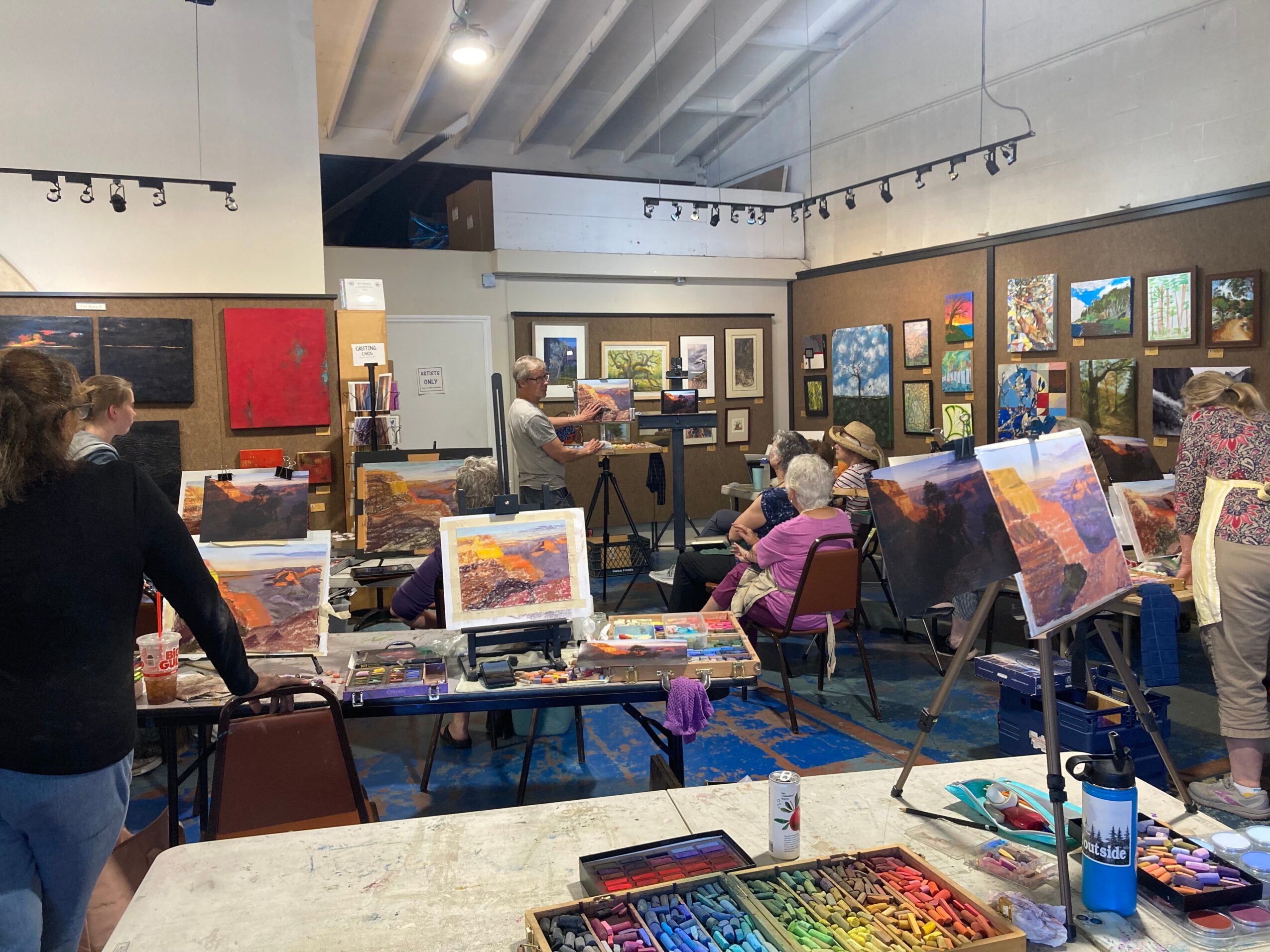 People in an art studio viewing and creating landscape paintings, with canvases on easels and art supplies on tables.