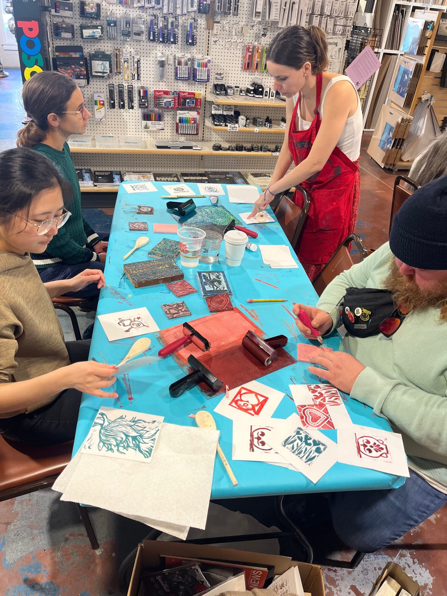 People gathered around a table with artwork, papers, and art supplies in a store or workshop setting.