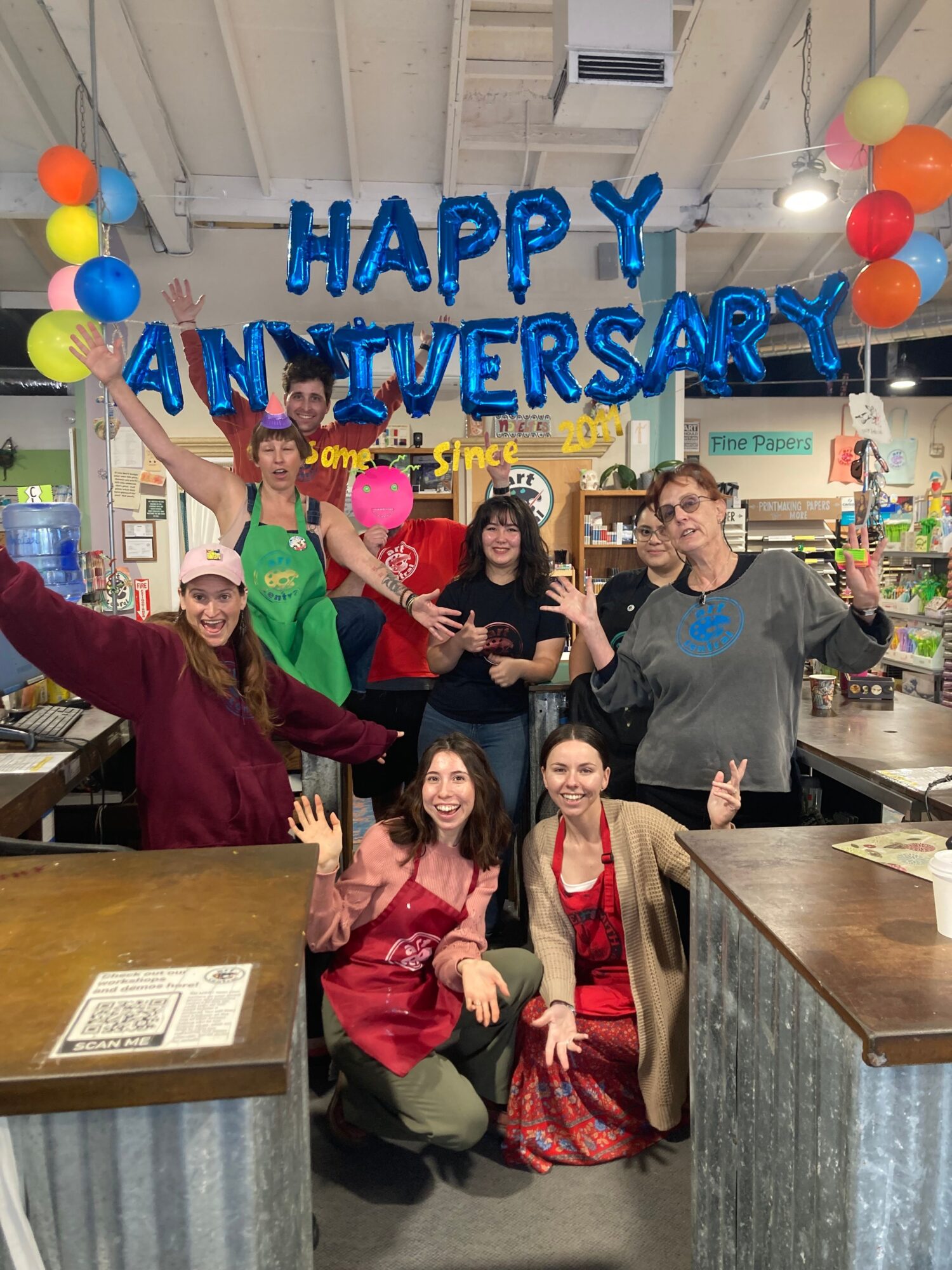 Group of five people celebrating an anniversary with balloons and decorations in a room, some sitting and some standing.