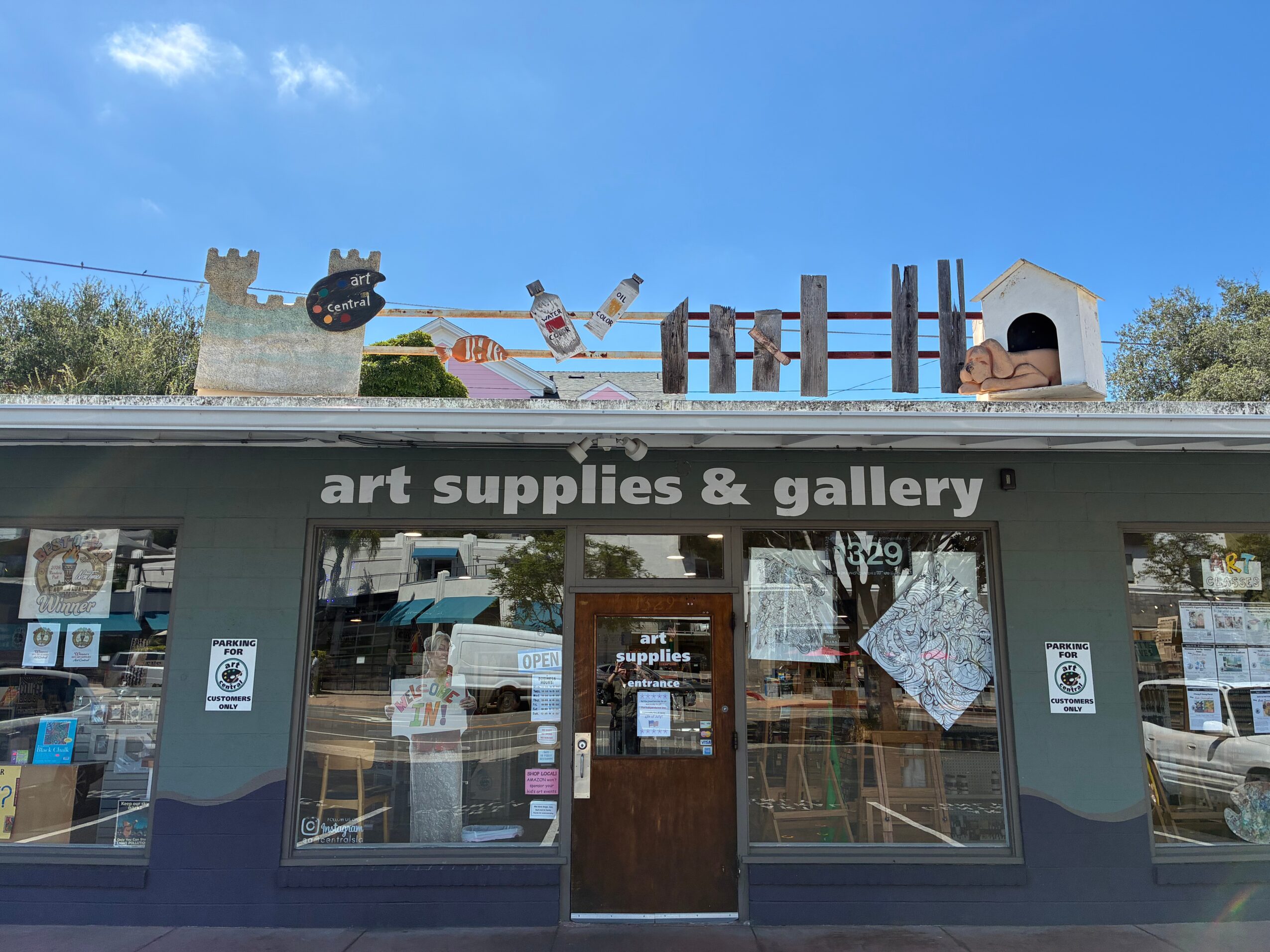 Storefront with art supplies and gallery sign, window displays, and a door, under a blue sky.