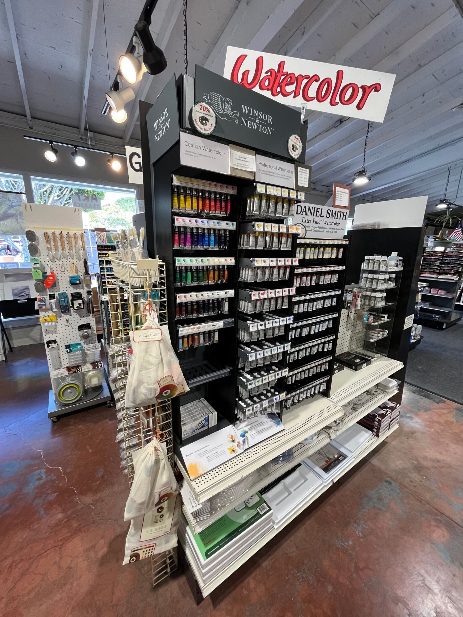 Display of watercolor art supplies including paints, brushes, and paper in a store, with a Watercolor sign visible.