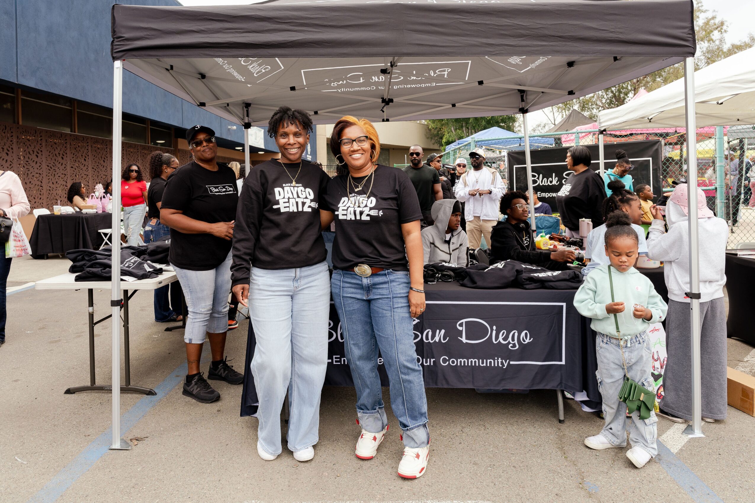 Two women stand in front of a booth at an outdoor event, smiling, with people and tents in the background.