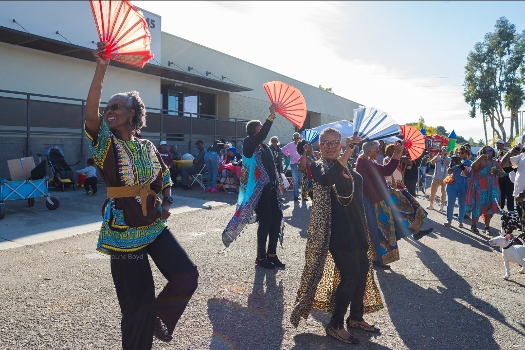 Group of people dancing outdoors with fans, some wearing colorful traditional clothing, in a sunny setting.