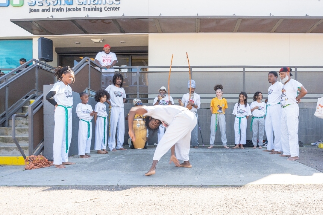 Group of children and adults watching a person perform a capoeira move outside a building with a sign reading 'Joan and Irwin Jacobs Training Center'.