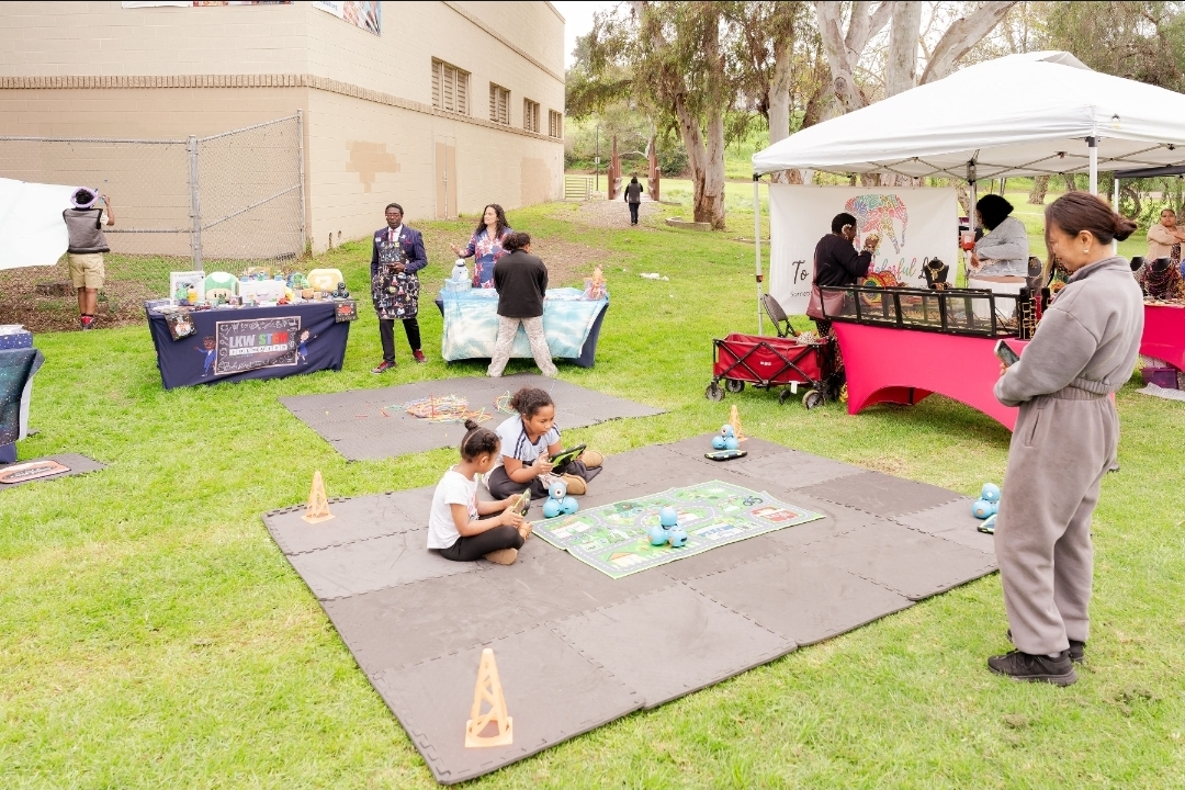 People at outdoor market stalls and children sitting on mats in a grassy area, with a tent and trees in the background.