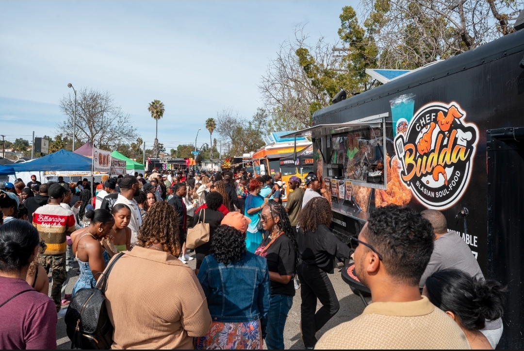 Crowd of people gathered outdoors near food trucks and tents, with trees and a clear sky in the background.