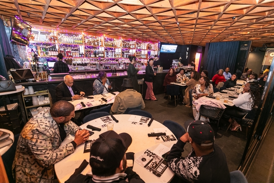 People dining and socializing in a restaurant with a bar in the background, warm lighting, and a wooden ceiling.