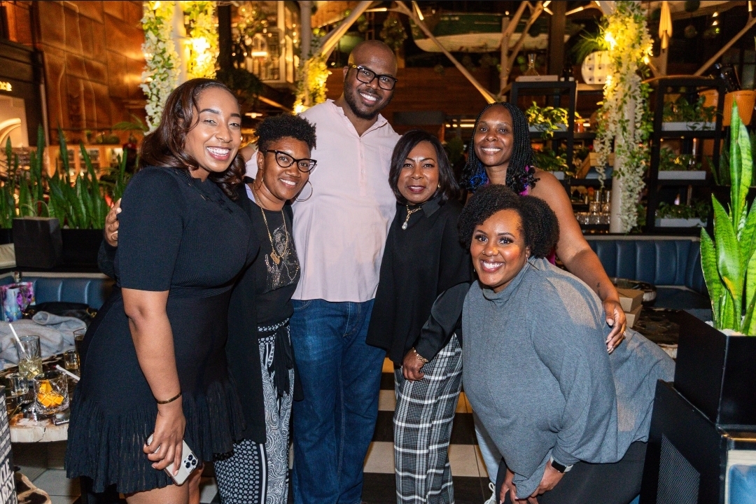 Group of six people smiling and posing together in a restaurant or bar setting with plants and warm lighting.