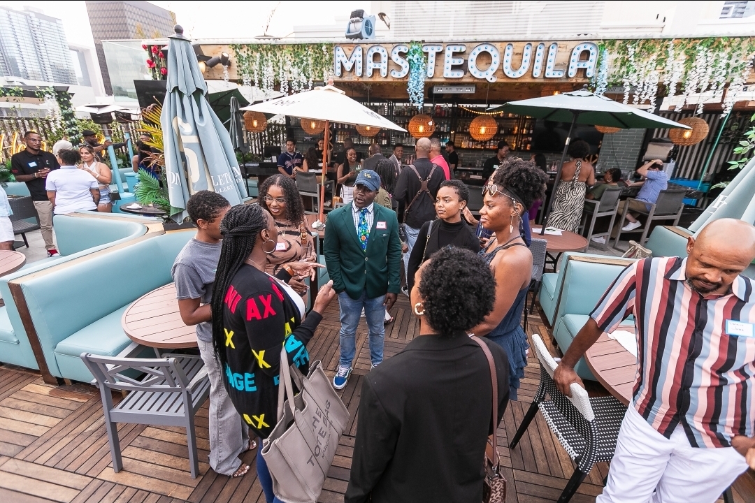 Group of people socializing outdoors at a bar with a sign reading 'MASTEEQUILA', surrounded by tables, umbrellas, and seating areas.