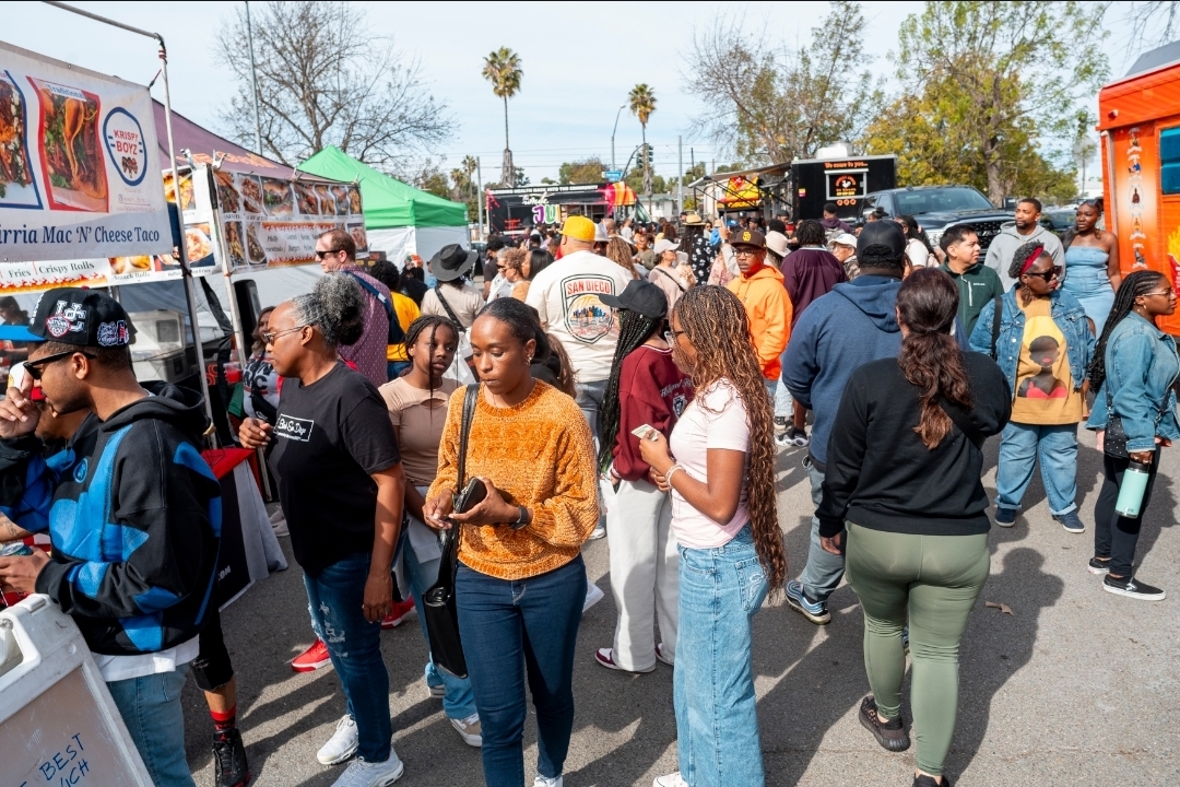 Crowd of people at an outdoor event with food trucks and tents, some holding food and drinks, trees in background.
