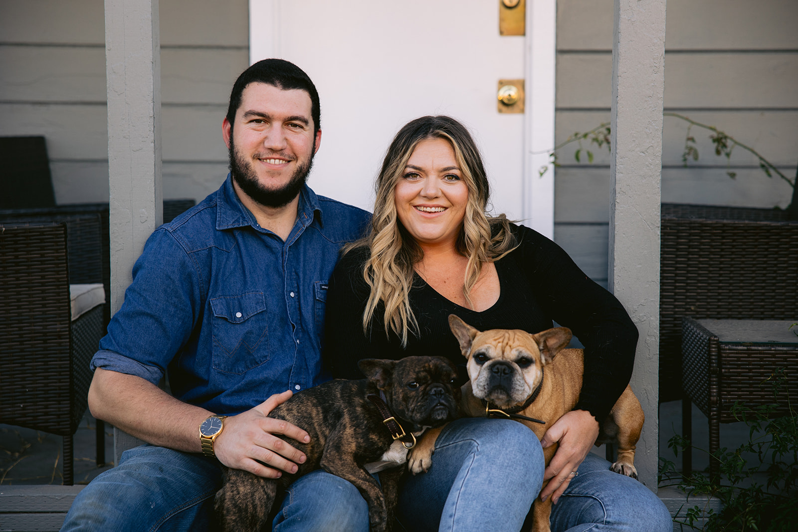A smiling couple sitting outdoors with two dogs, one small and one large, in front of a gray wooden wall.