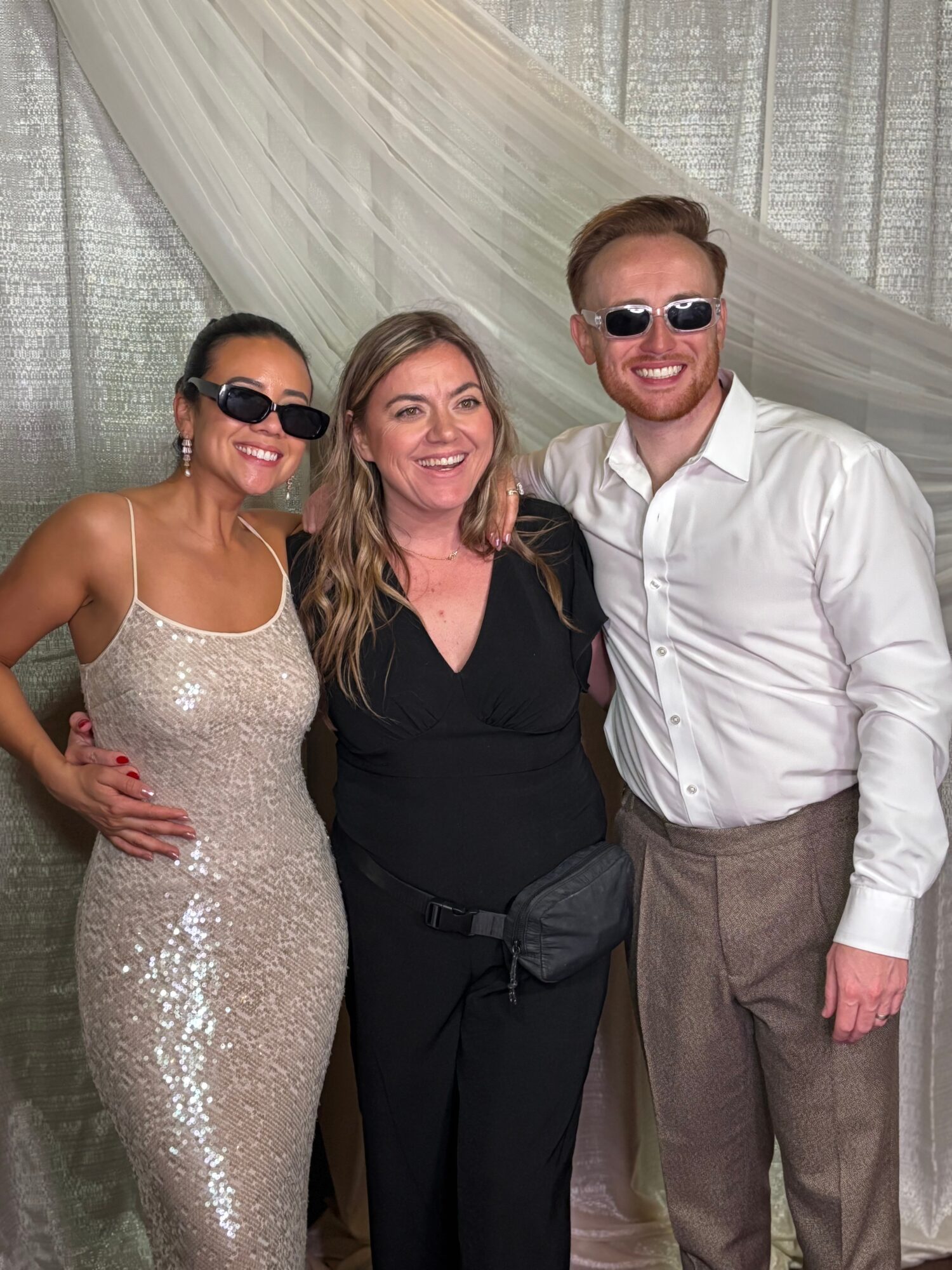 Three people smiling, standing close together indoors with a draped curtain background.