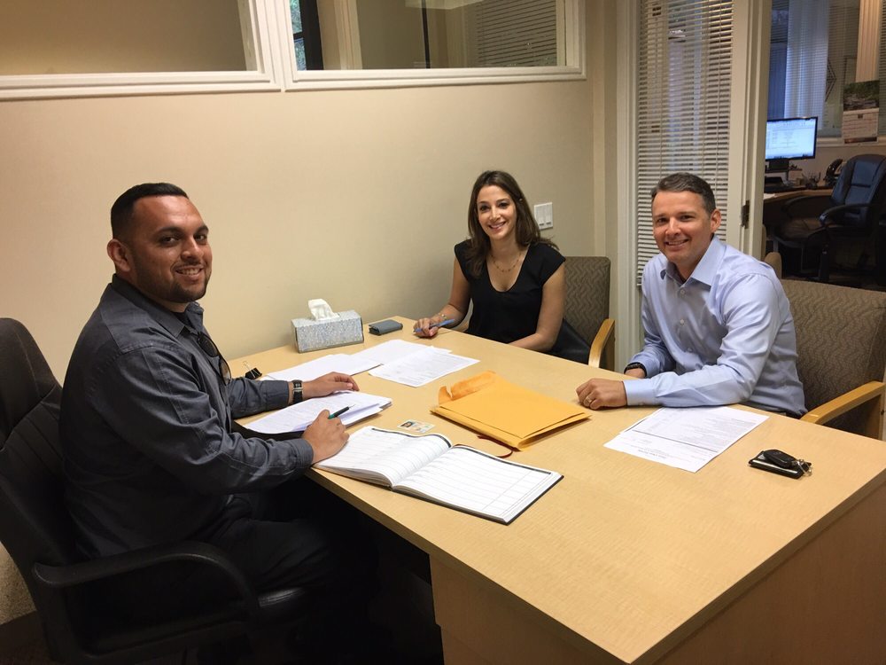 Three people sitting at a desk in an office, smiling, with papers and folders on the desk.