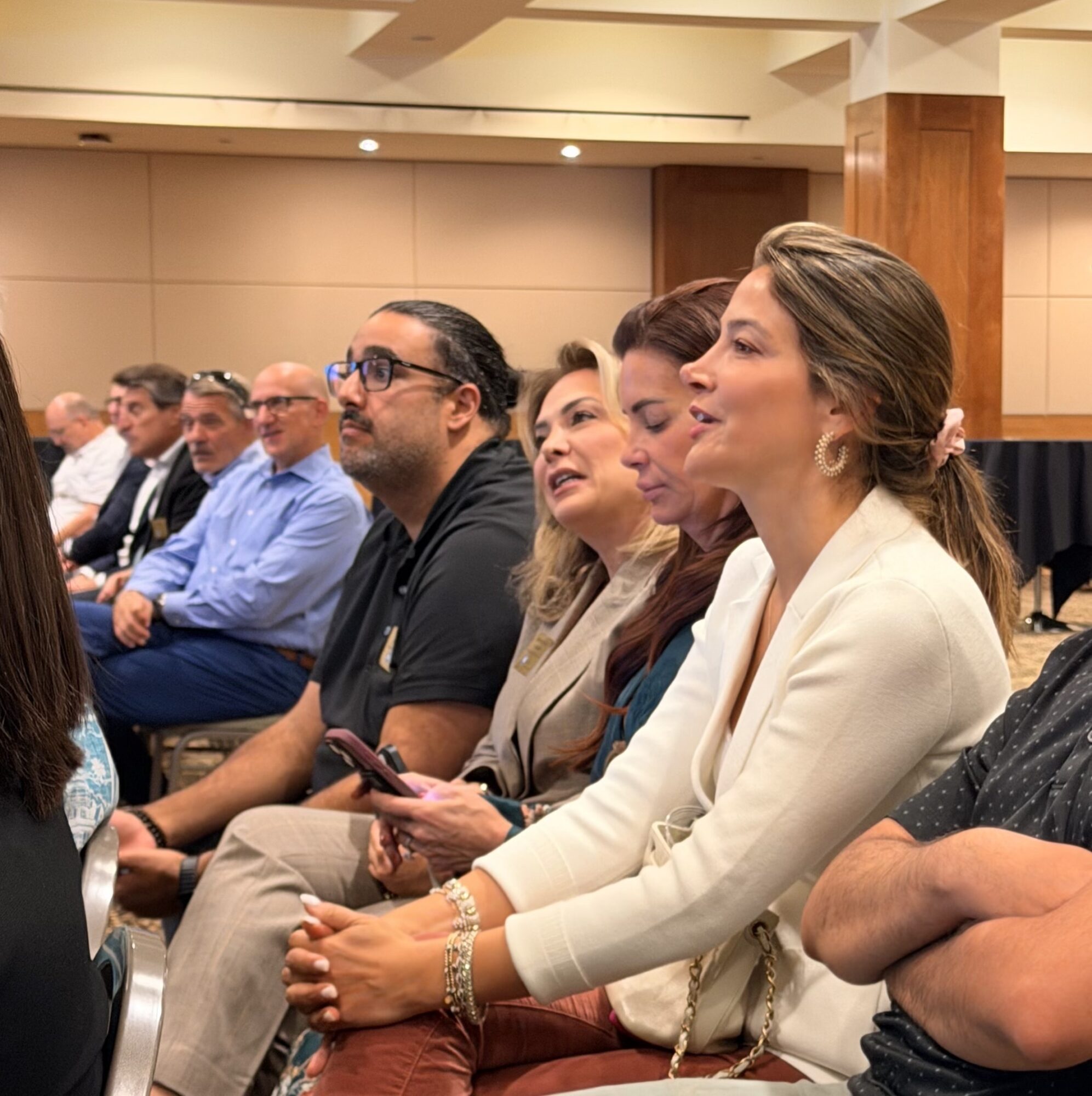 Group of people seated in a conference room, listening attentively, with a woman in white in the foreground smiling.