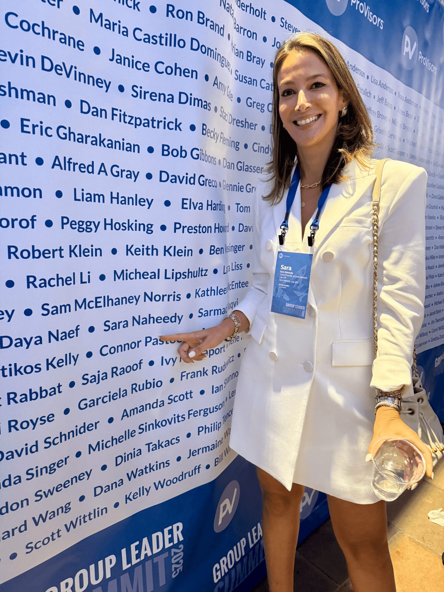 Smiling woman in white dress and blazer standing next to a wall with names and logos, holding a phone and a bag.