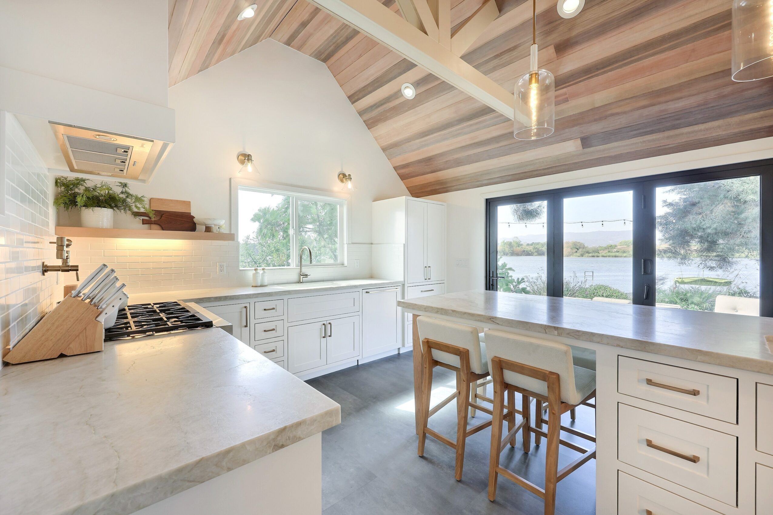 Kitchen with white cabinets, a large window, and a wooden ceiling with pendant lights, island with two chairs, and a view outside.