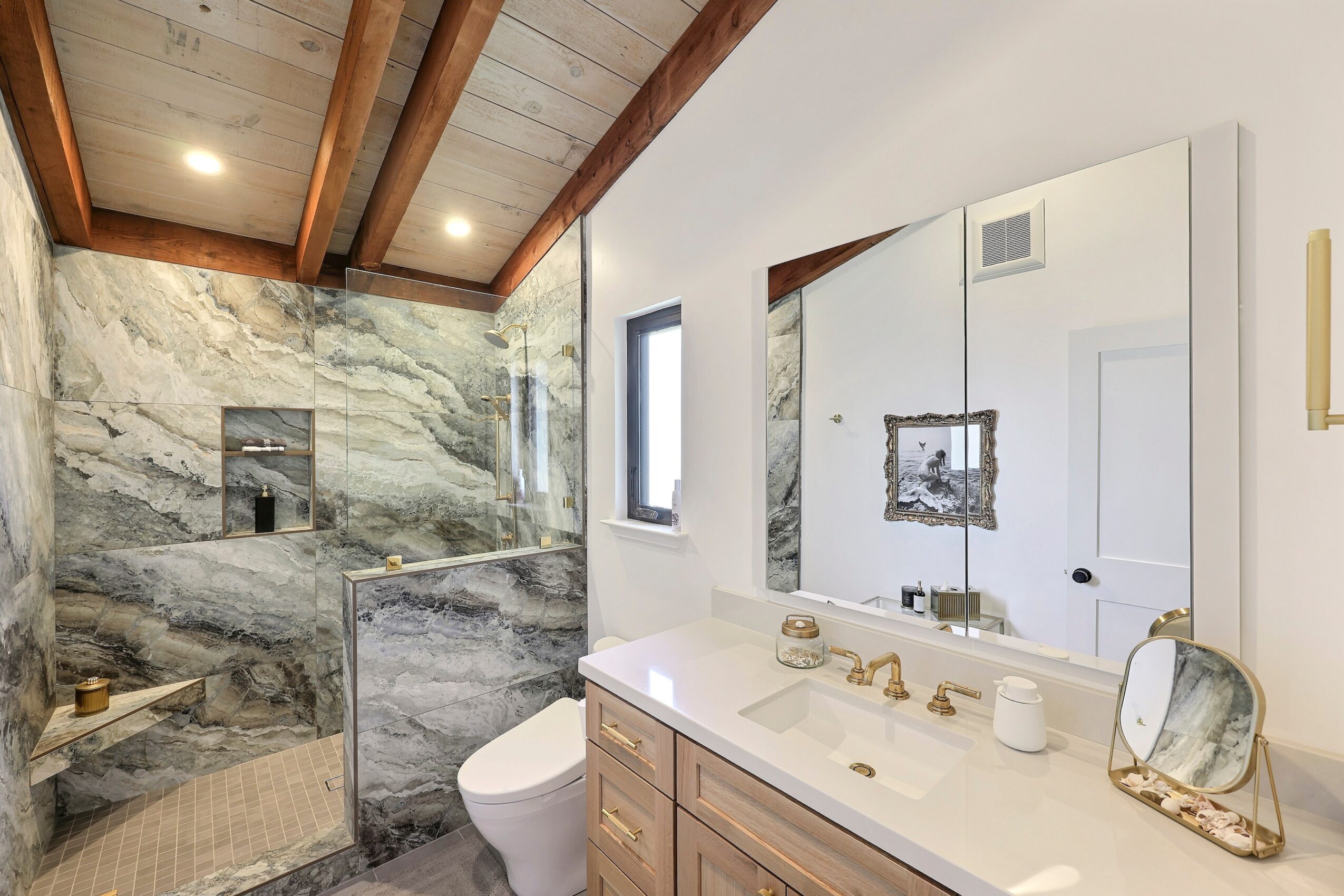 Bathroom with marble walls, wooden ceiling, white vanity, mirror, and small window, featuring modern fixtures and decor.