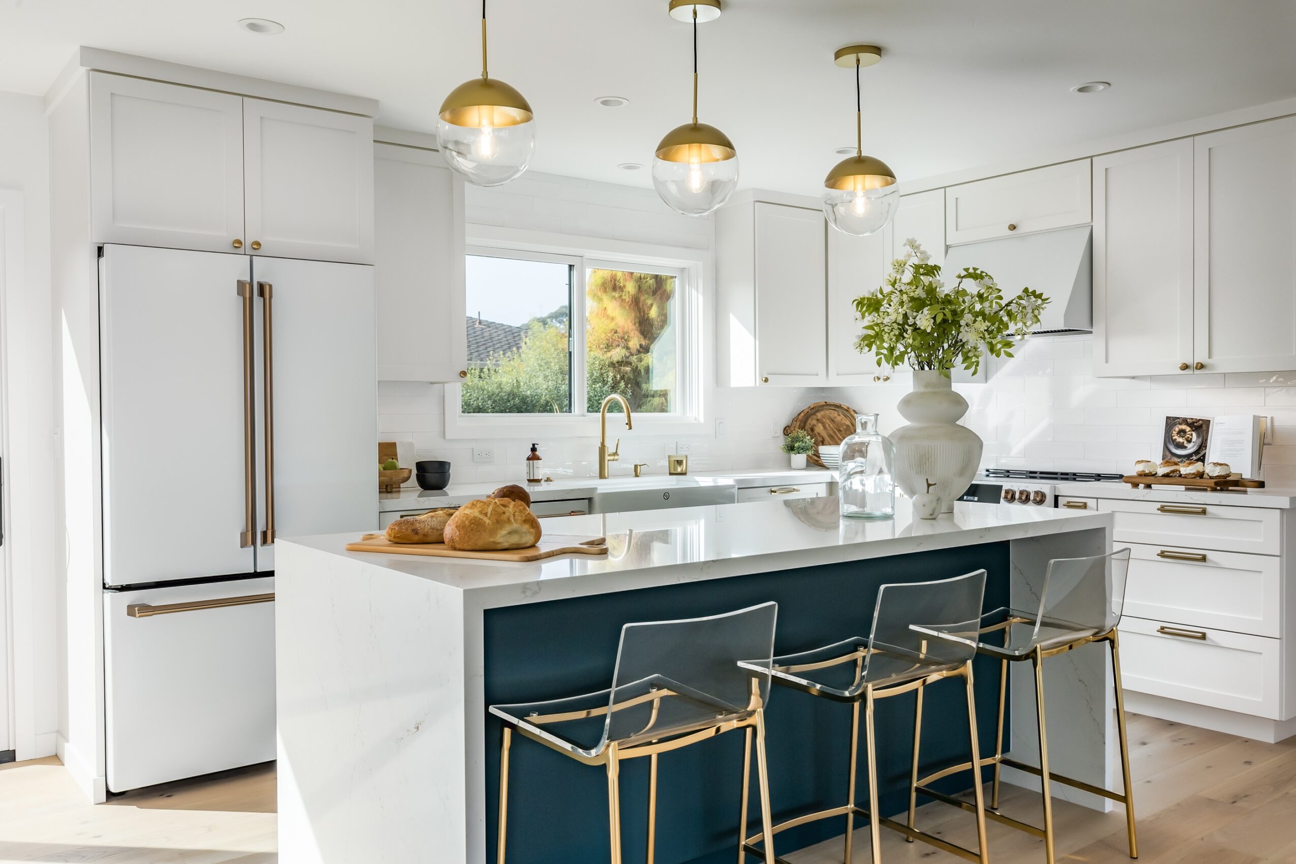 Modern kitchen with white cabinets, island, three gold pendant lights, and a window with greenery outside.