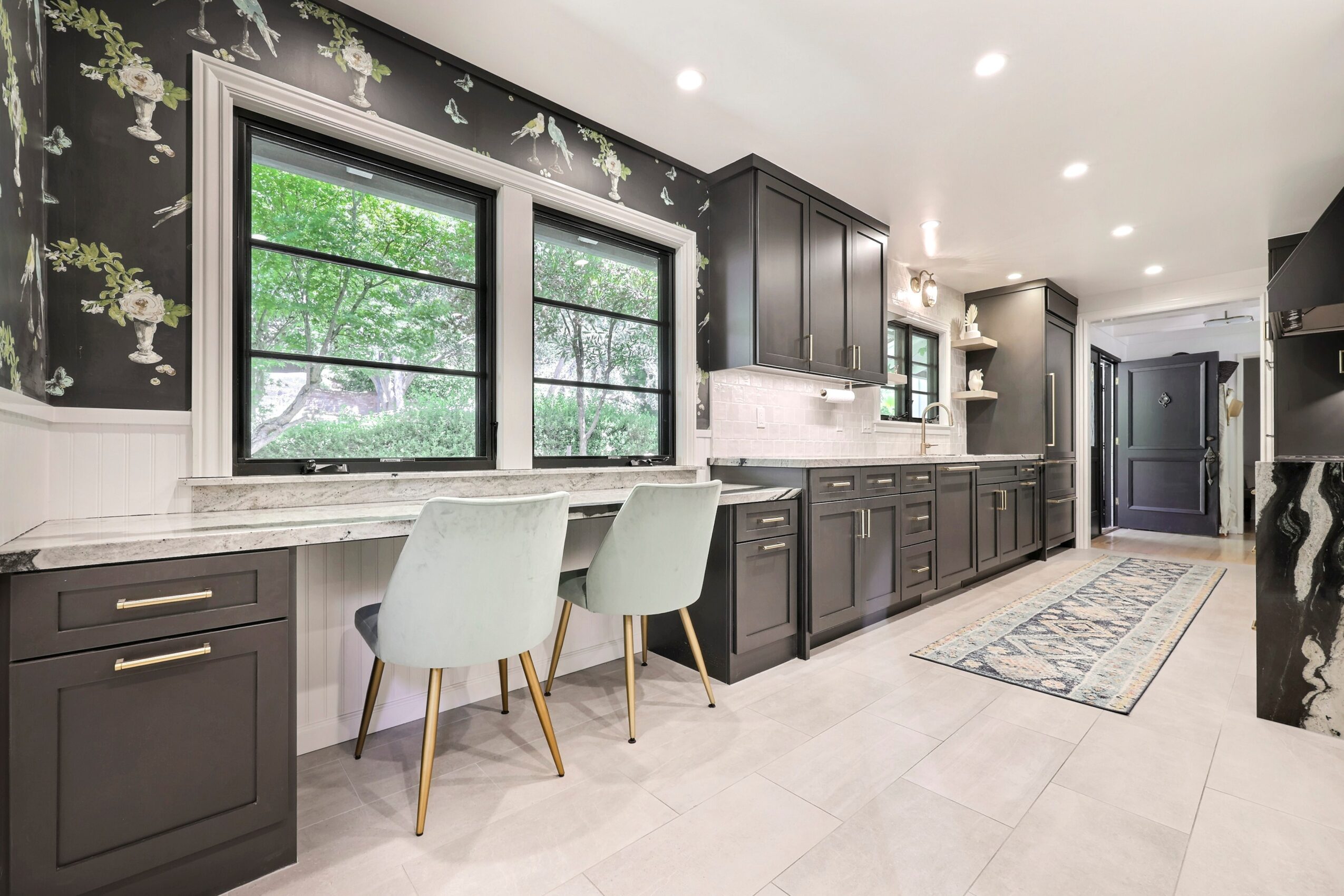 Kitchen with black cabinets, white countertops, two white chairs by window, patterned wallpaper, and a rug on light-colored floor.