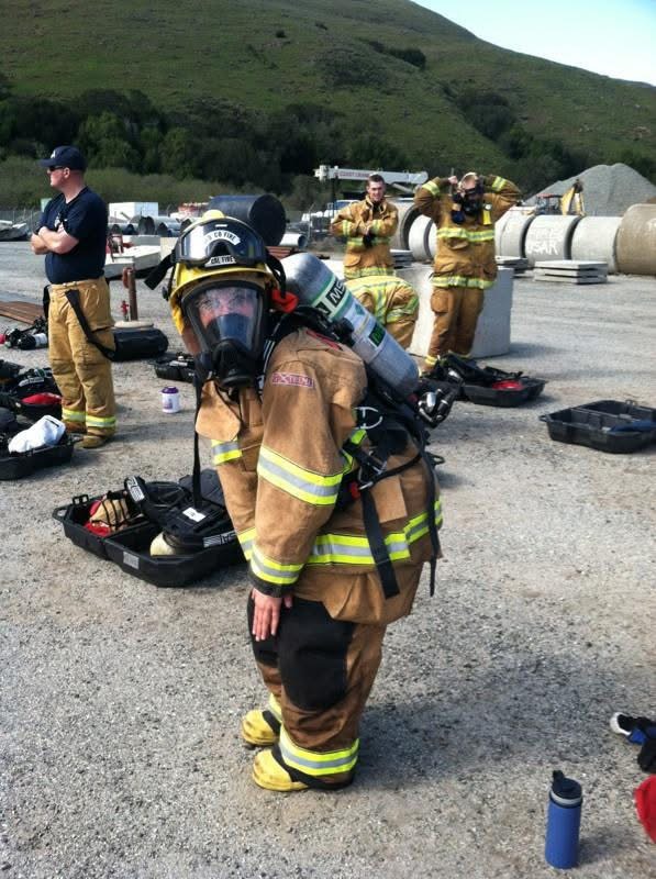 Firefighter in full gear kneeling on the ground with other firefighters and equipment in the background outdoors.