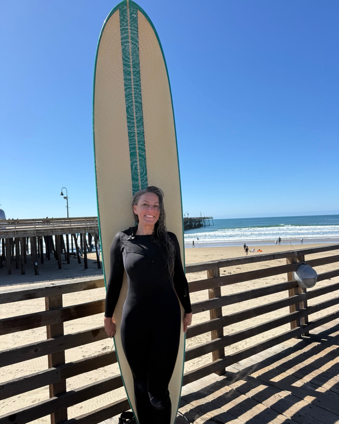 Woman standing on a beach boardwalk holding a surfboard, with ocean and sky in background.