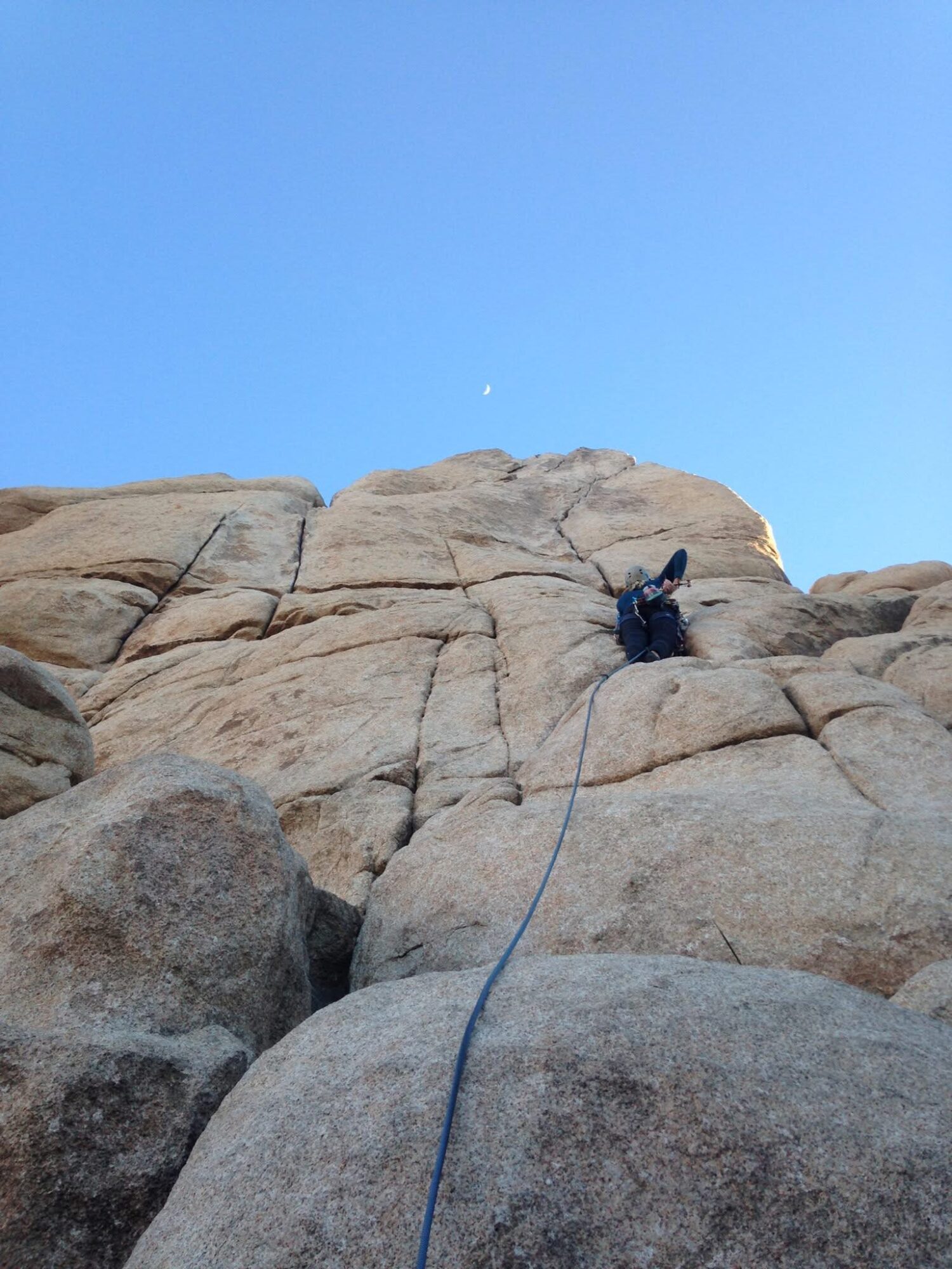 Person climbing large rock formation with a rope, clear blue sky above, small moon visible, rocky terrain.