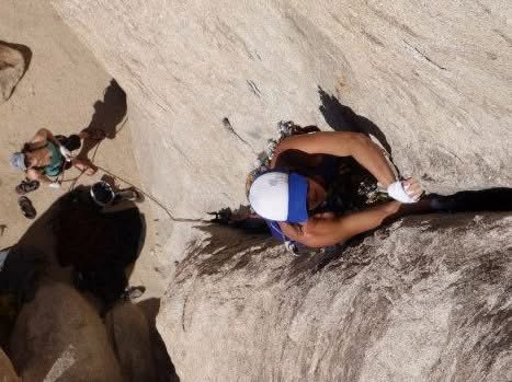 Person climbing a steep rock face with a harness and helmet, with another person holding the rope nearby.