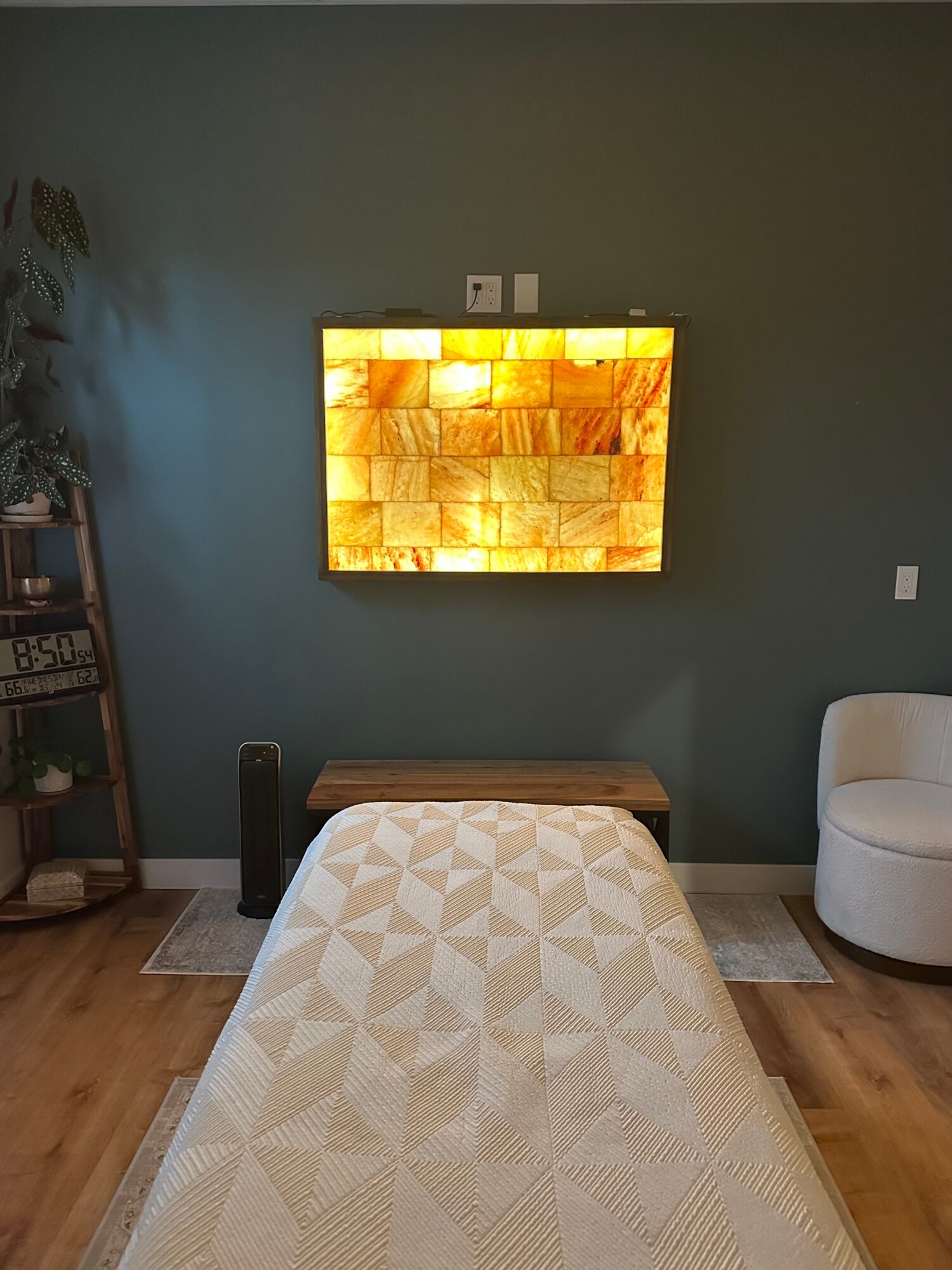 A room with a wall-mounted TV, a bed, a white armchair, and a small table, with a plant on the left.