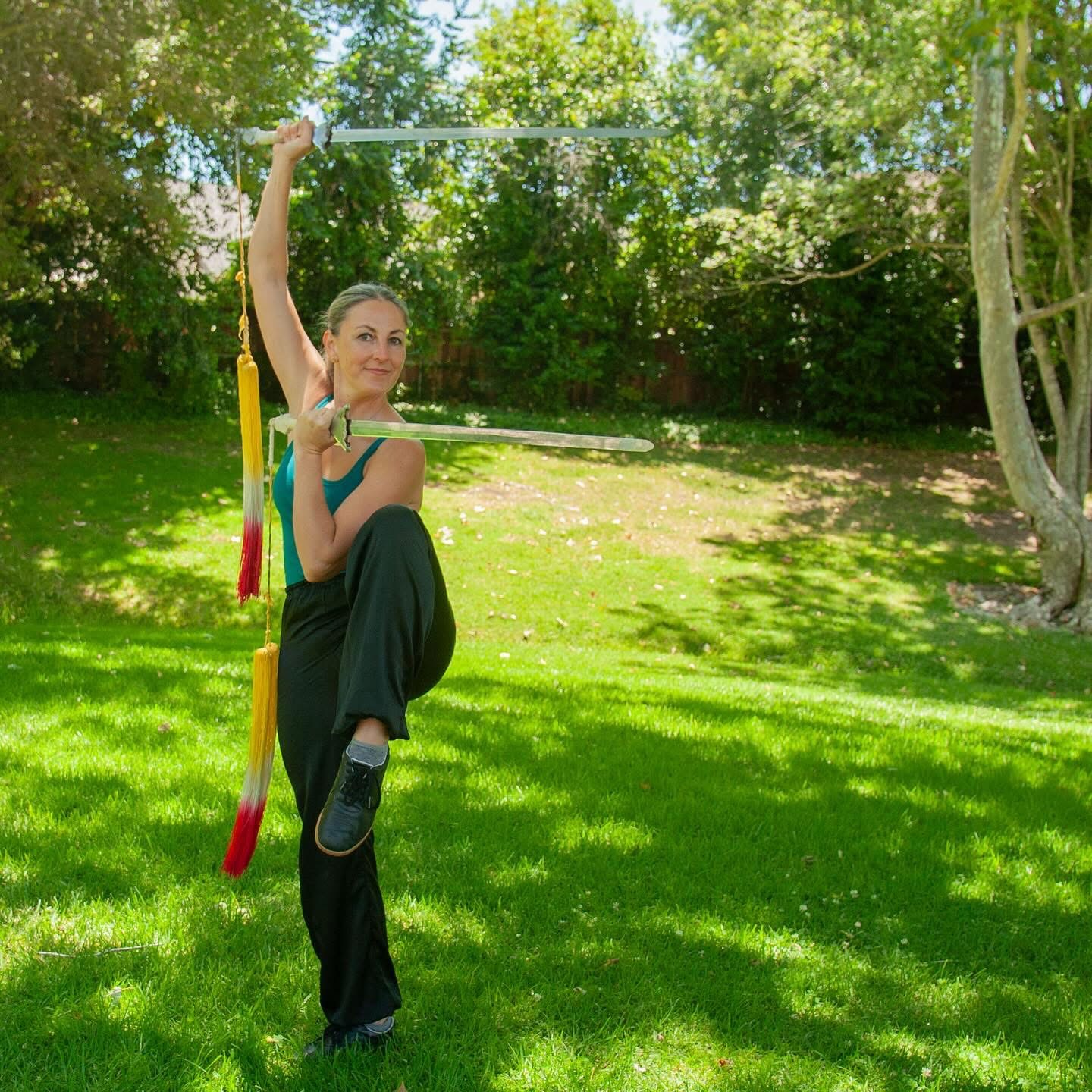 Woman practicing pole dance outdoors on grass, holding pole with one arm, one leg raised, surrounded by trees and sunlight.