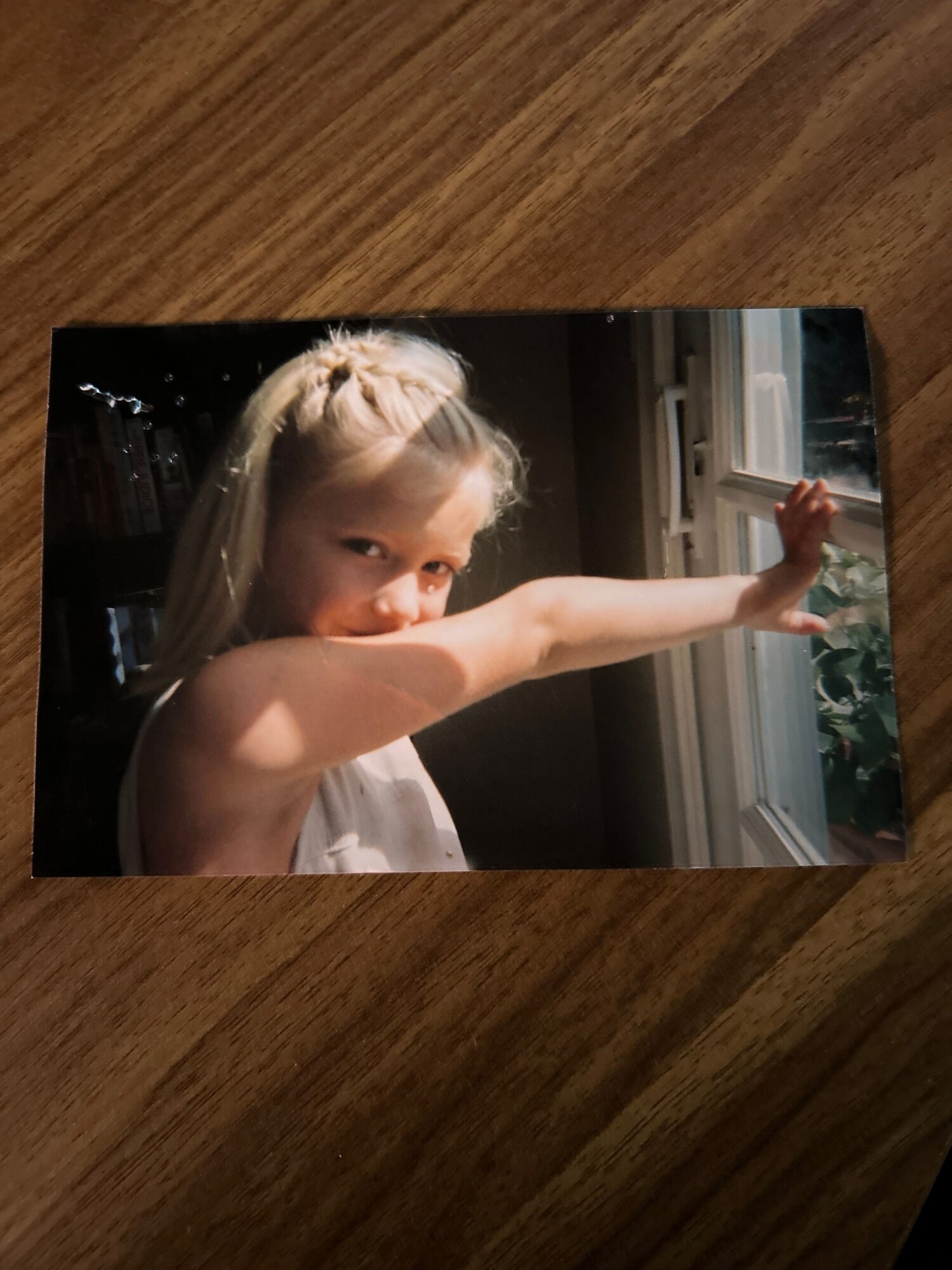 Child with blonde hair lying down, reaching upward with one arm, indoors with window in background.