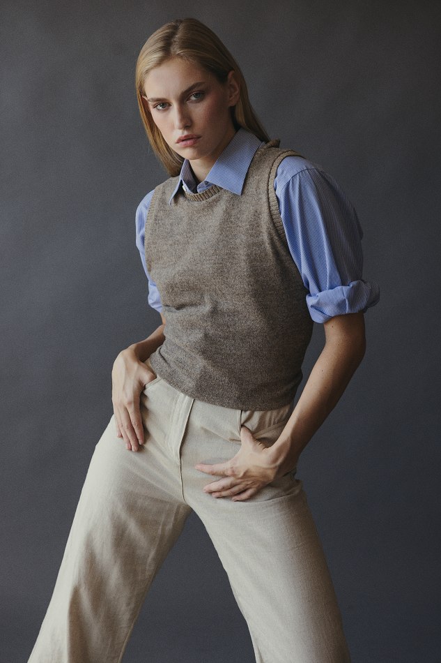 Young woman with blonde hair wearing a blue shirt, gray vest, and beige pants, posing against a dark background.