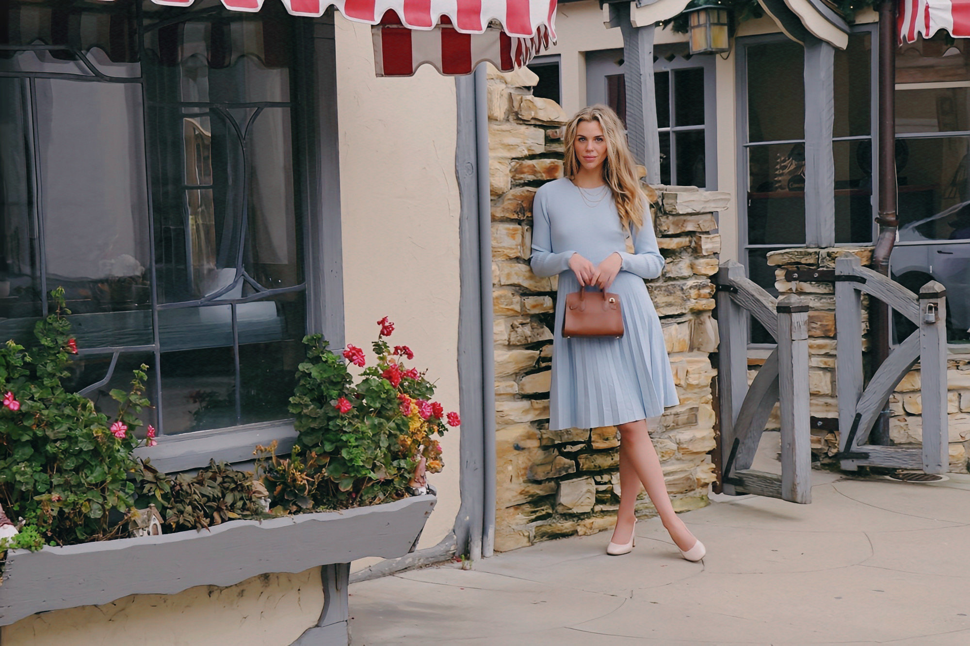 Woman in a light blue dress holding a brown bag, standing outside near a stone wall and flowers.