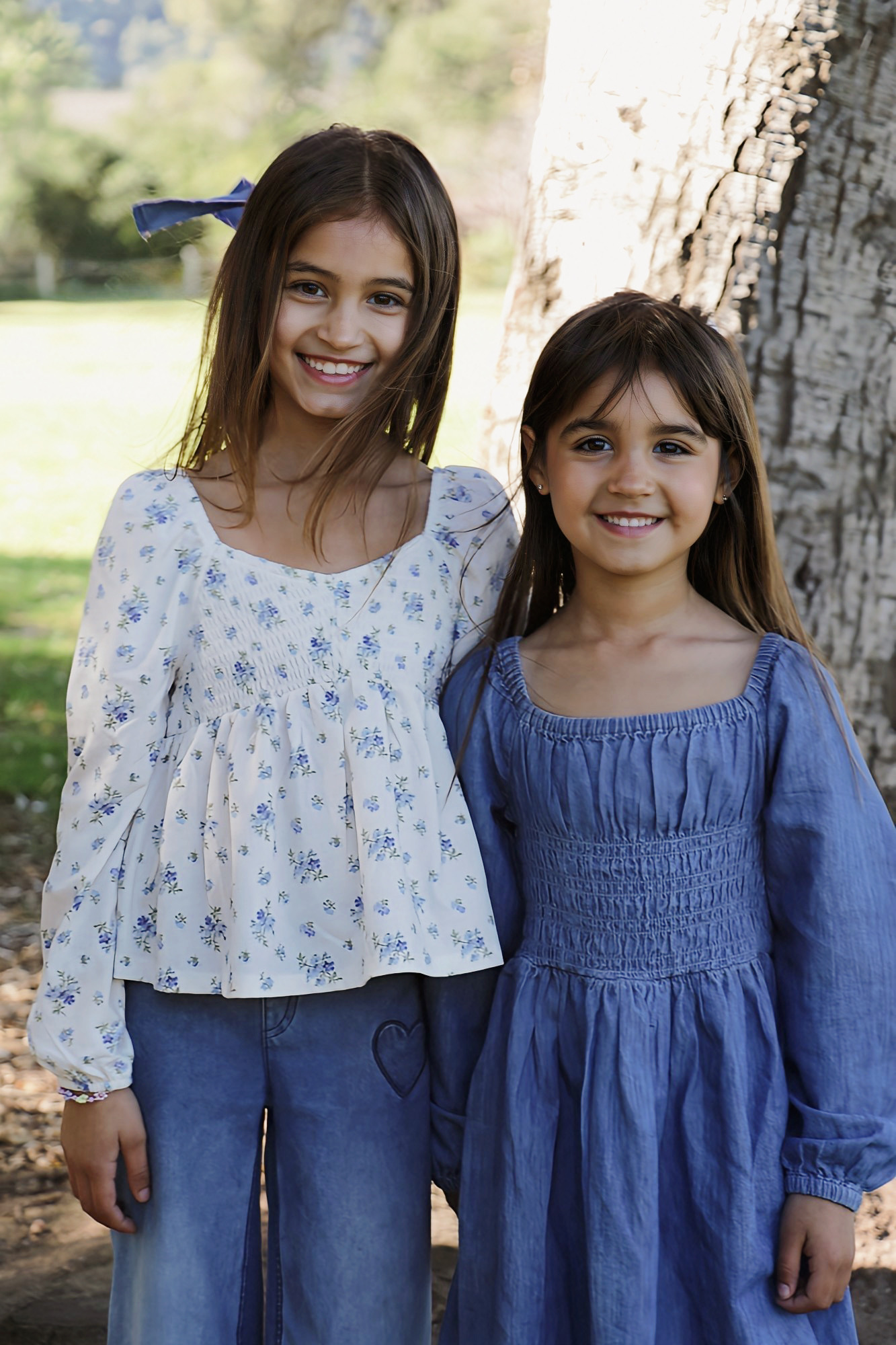 Two smiling girls stand outdoors near a tree, wearing blue dresses, with a grassy background and sunlight filtering through leaves.
