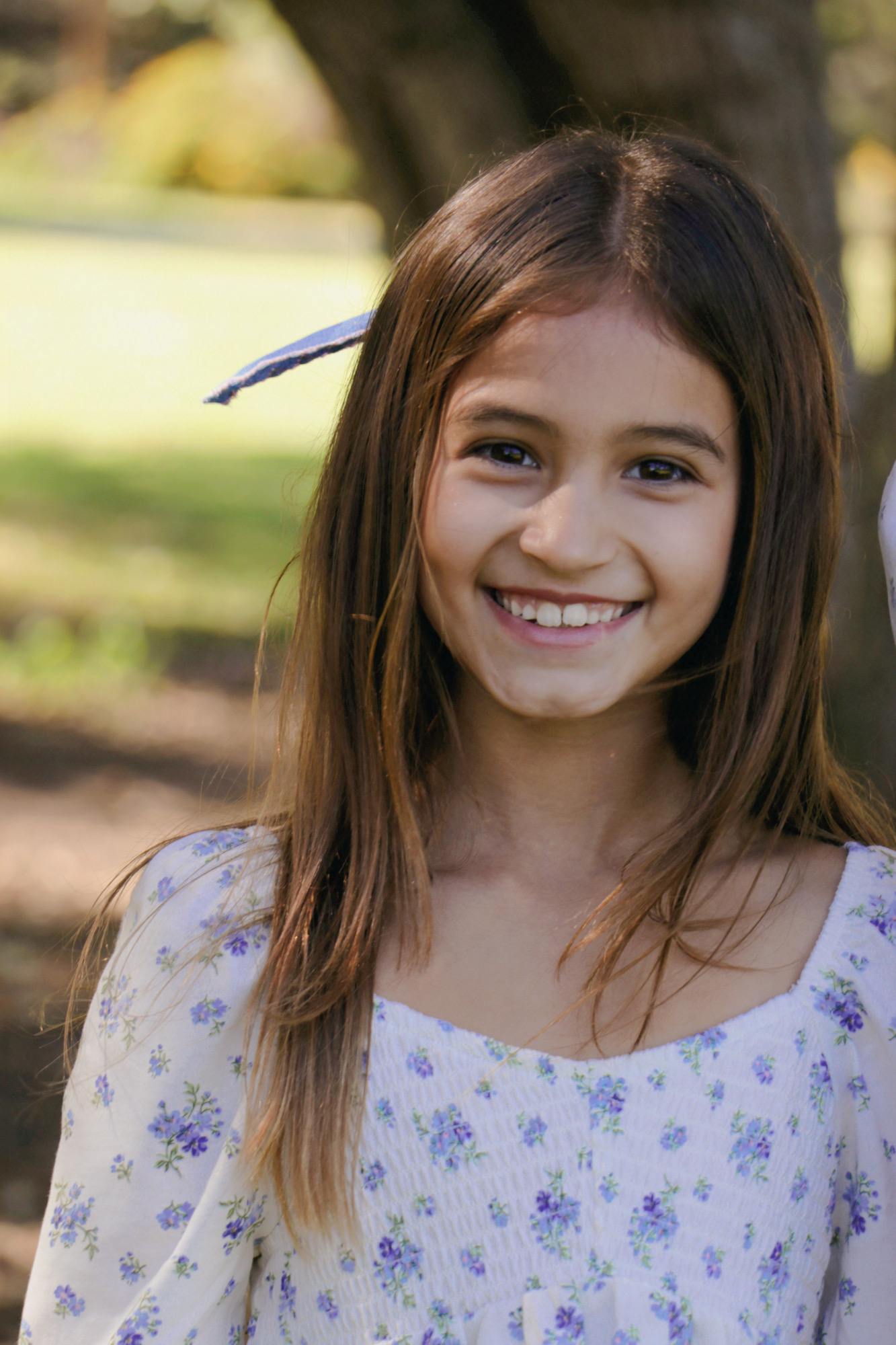 Young girl with long brown hair smiling outdoors, wearing a white floral dress, in a park setting.