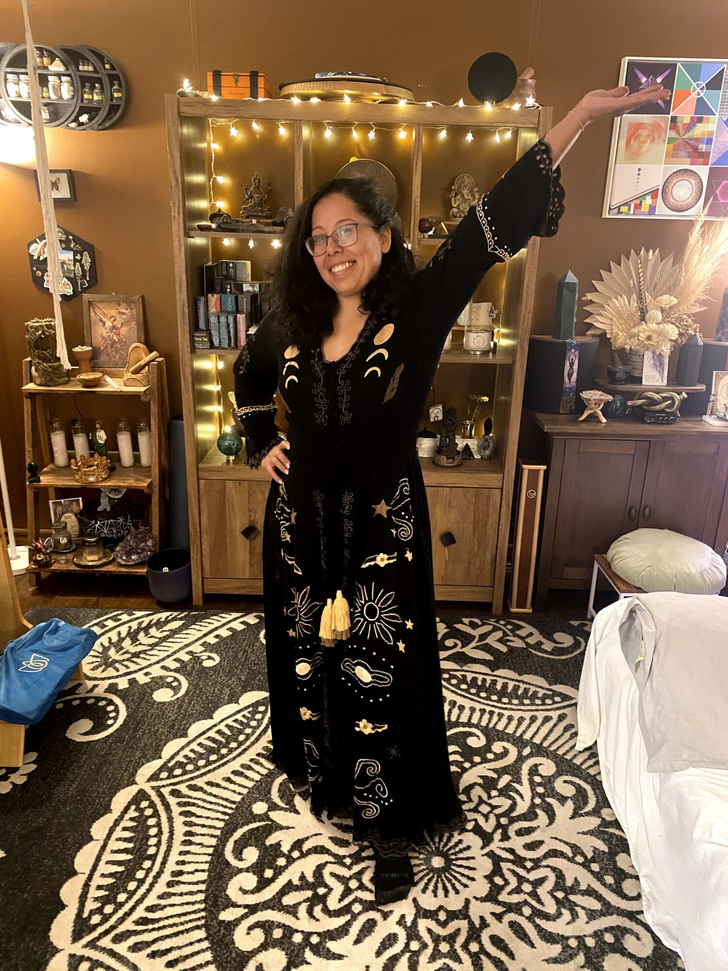Woman standing in a room with shelves, fairy lights, and a patterned rug, raising her arm and smiling.