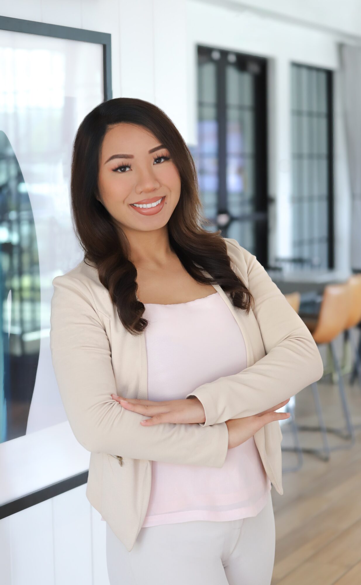 Smiling woman with long dark hair, wearing a light-colored blazer and top, standing indoors with large windows behind her.