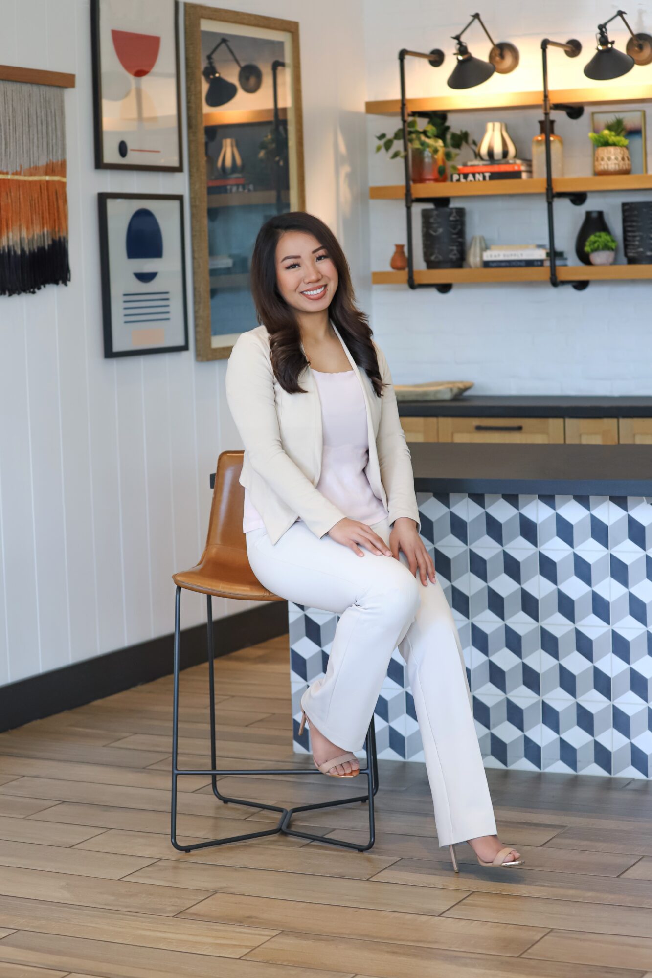 Woman sitting on a barstool in a modern interior with artwork and shelves, smiling, wearing white outfit.