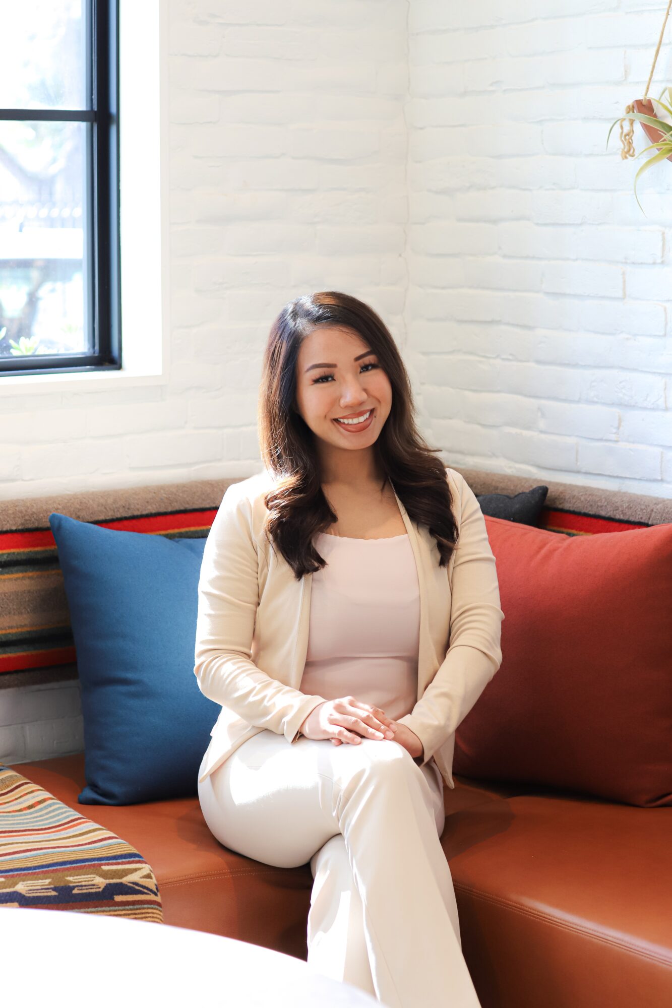 Smiling woman sitting on a sofa with colorful pillows in a bright room with a window and white brick wall.