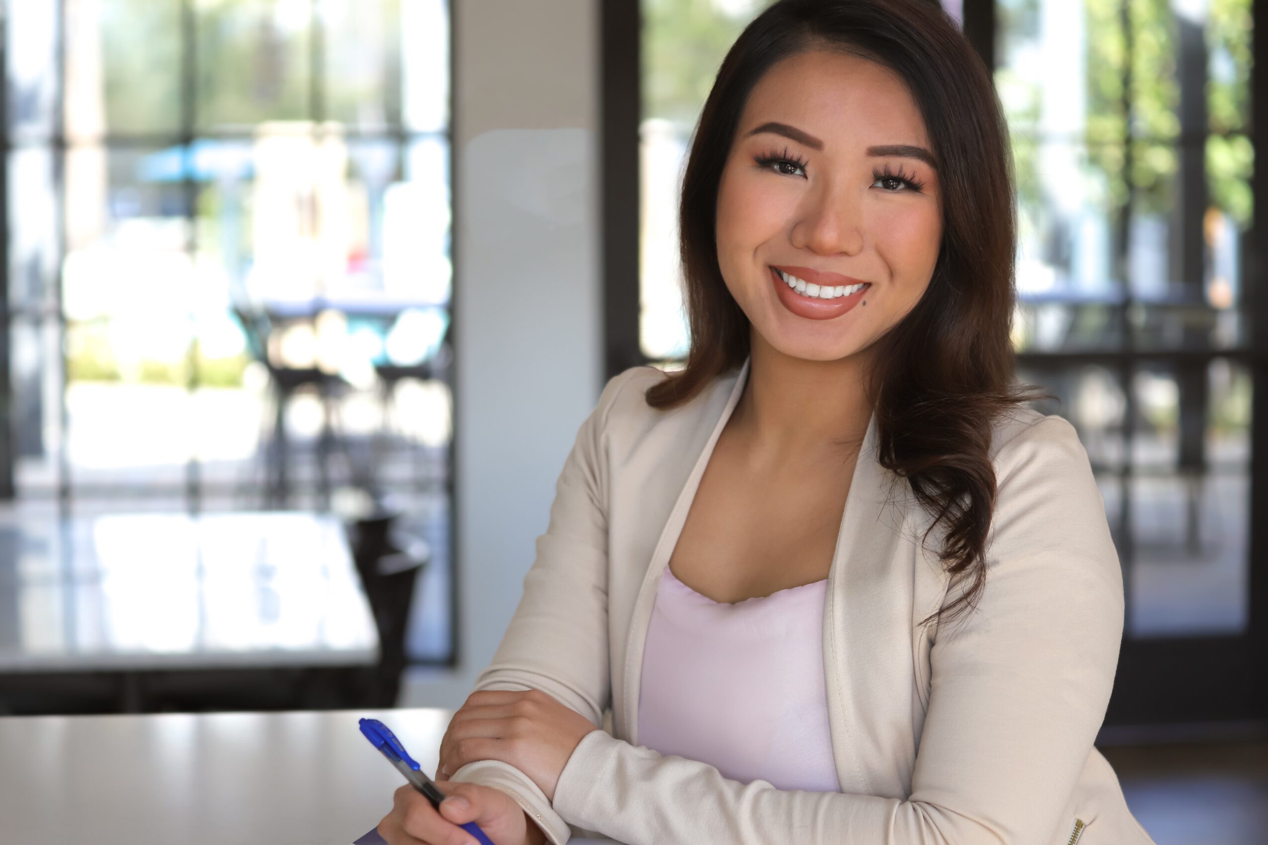 Smiling woman with long dark hair sitting indoors, background shows large windows and outdoor scenery.