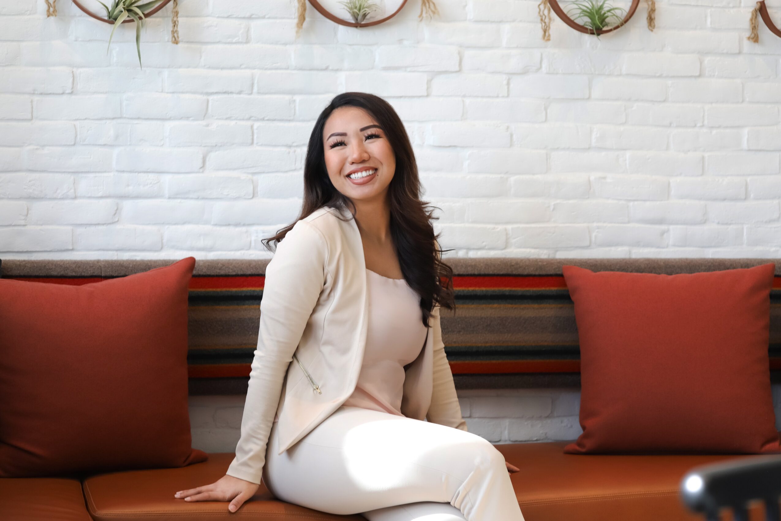 Smiling woman sitting on a red couch with a white brick wall background, wearing a beige blazer and white pants.