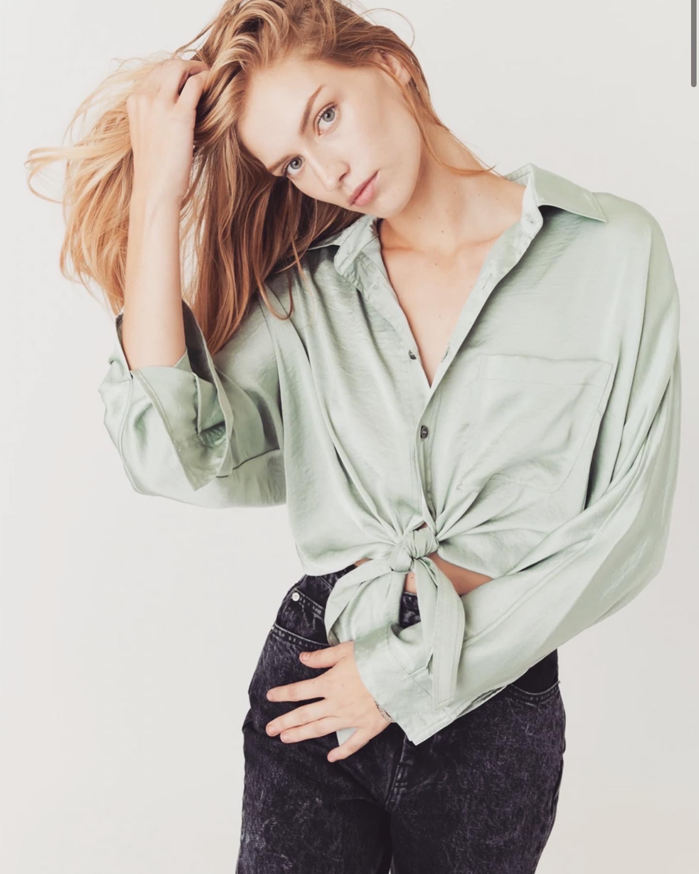 Young woman with blonde hair wearing a light green shirt tied at the waist, posing with her hand in her hair.
