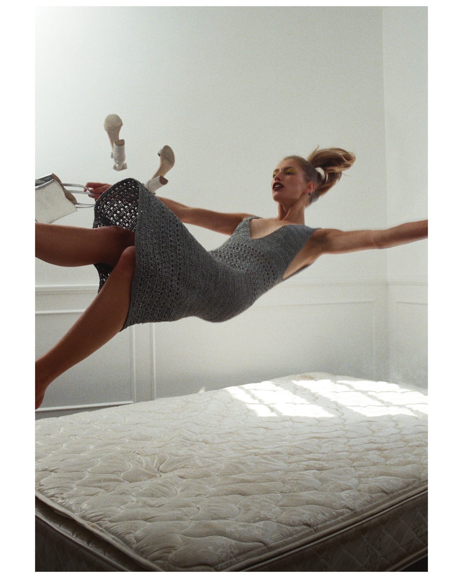Person mid-air above a bed, arms and legs extended, smiling, in a room with white walls and sunlight.