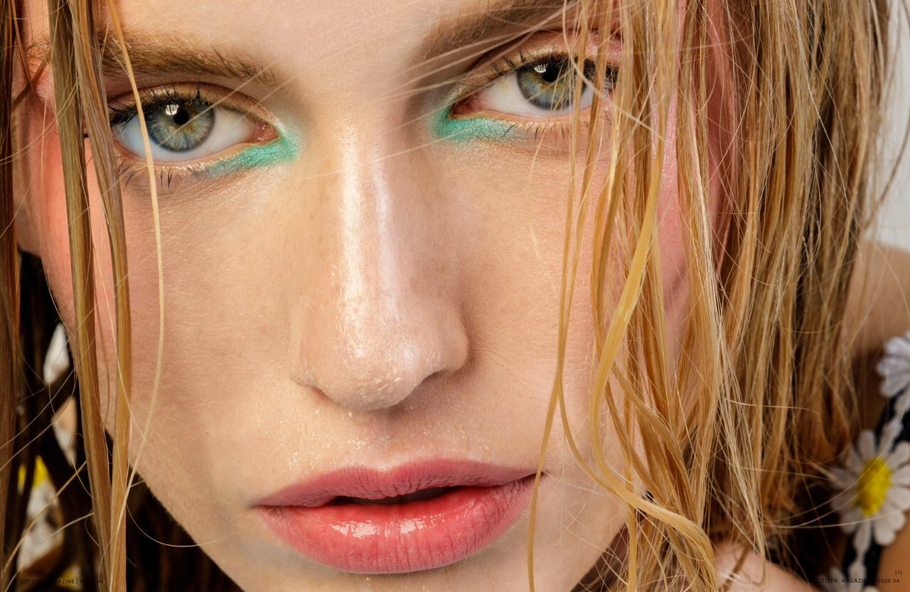 Close-up of a woman's face with blue eyes and light makeup, wet hair, and a daisy on her shoulder.