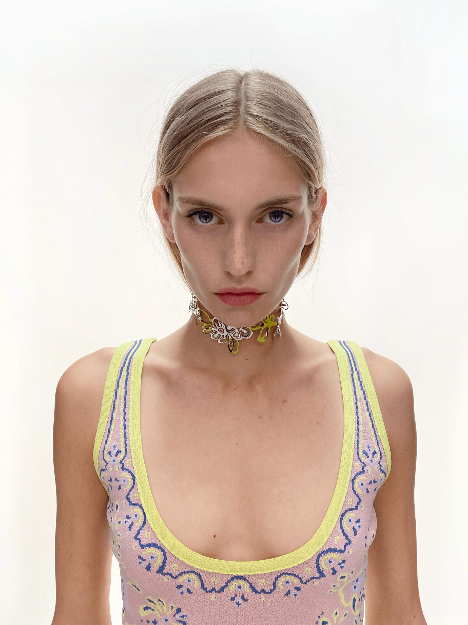 Young woman with blonde hair in a tank top, wearing a floral choker necklace, facing forward with a neutral expression.