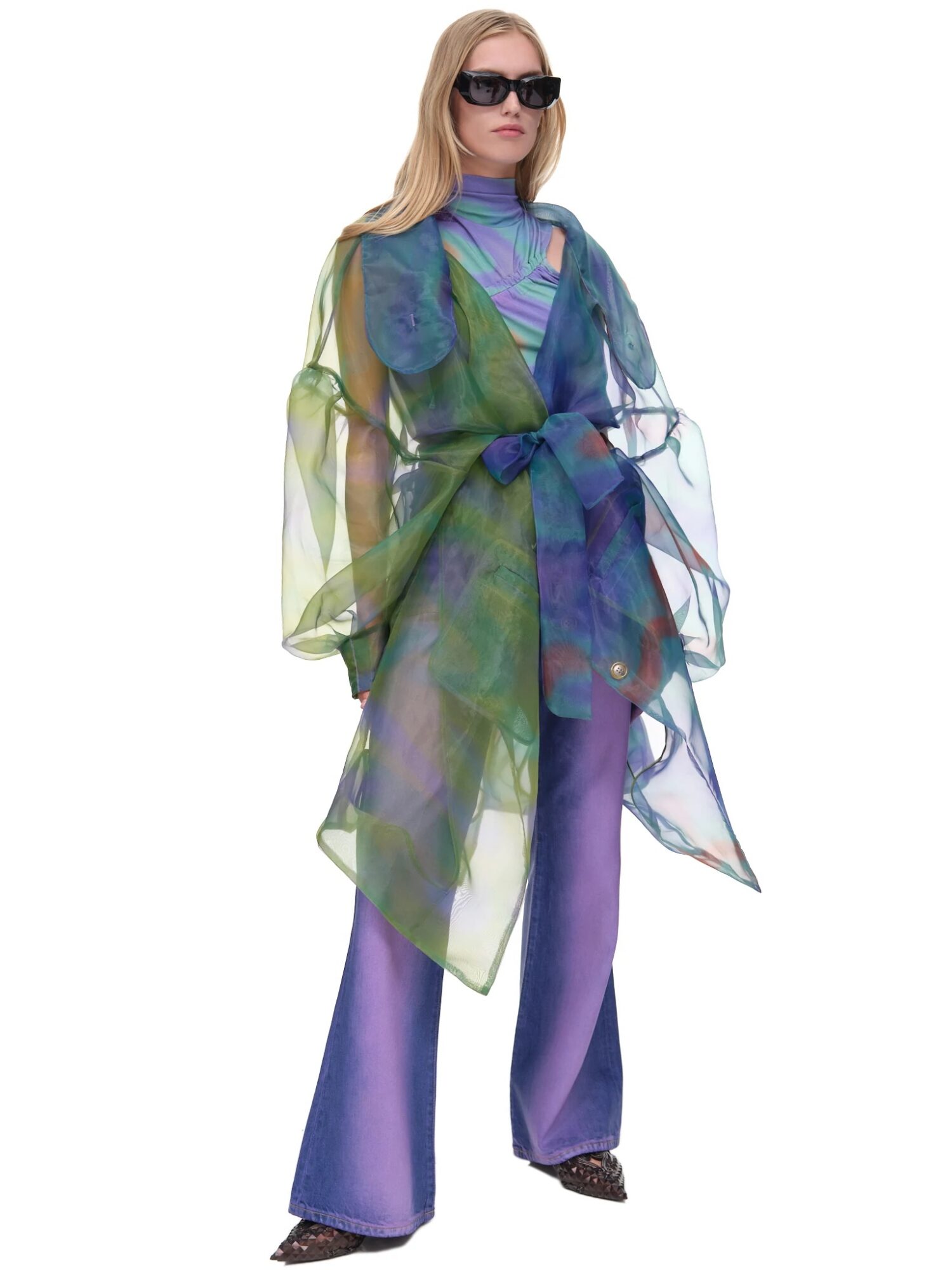 Woman wearing sunglasses, a colorful sheer top over a dress, and wide-leg pants, standing against a white background.