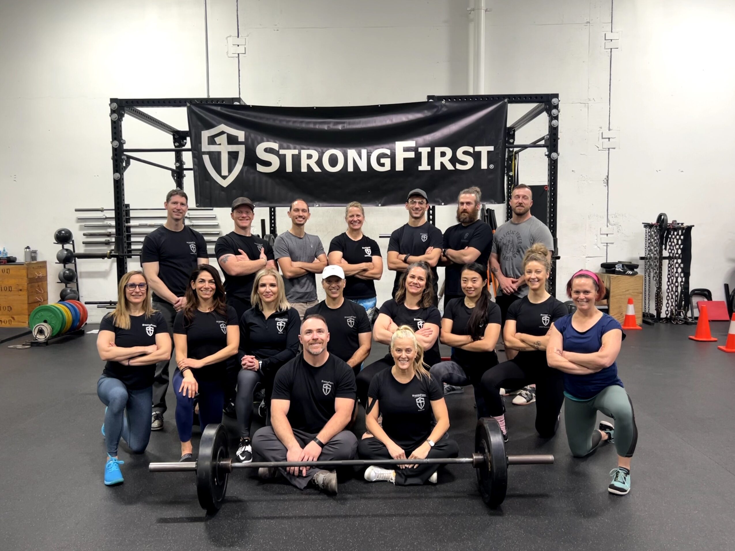 Group of people in a gym with a banner reading 'StrongFirst', some with arms crossed, standing behind a barbell on the floor.