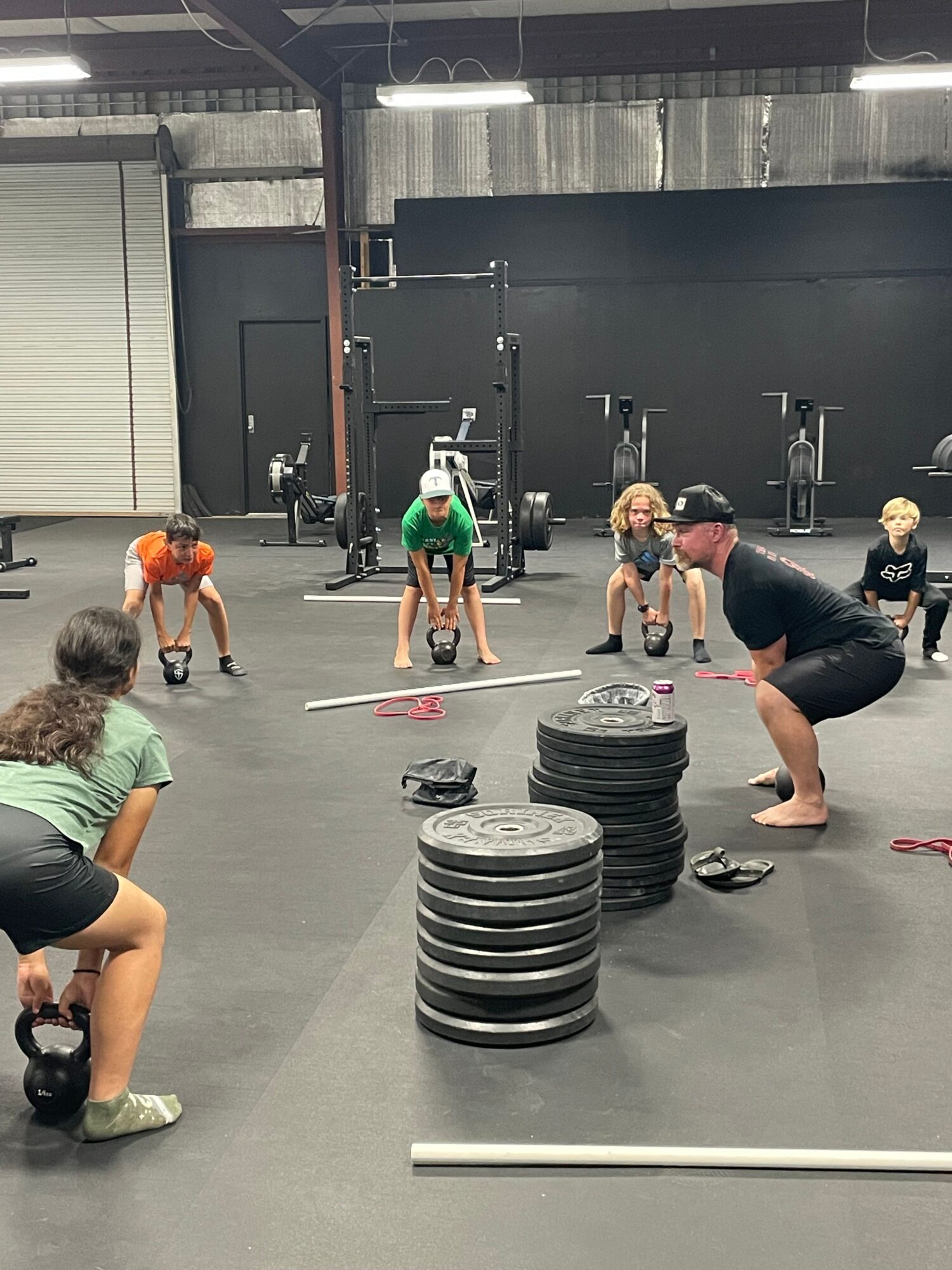 People exercising in a gym with weights and equipment, some lifting kettlebells, others standing or bending, in a spacious room.