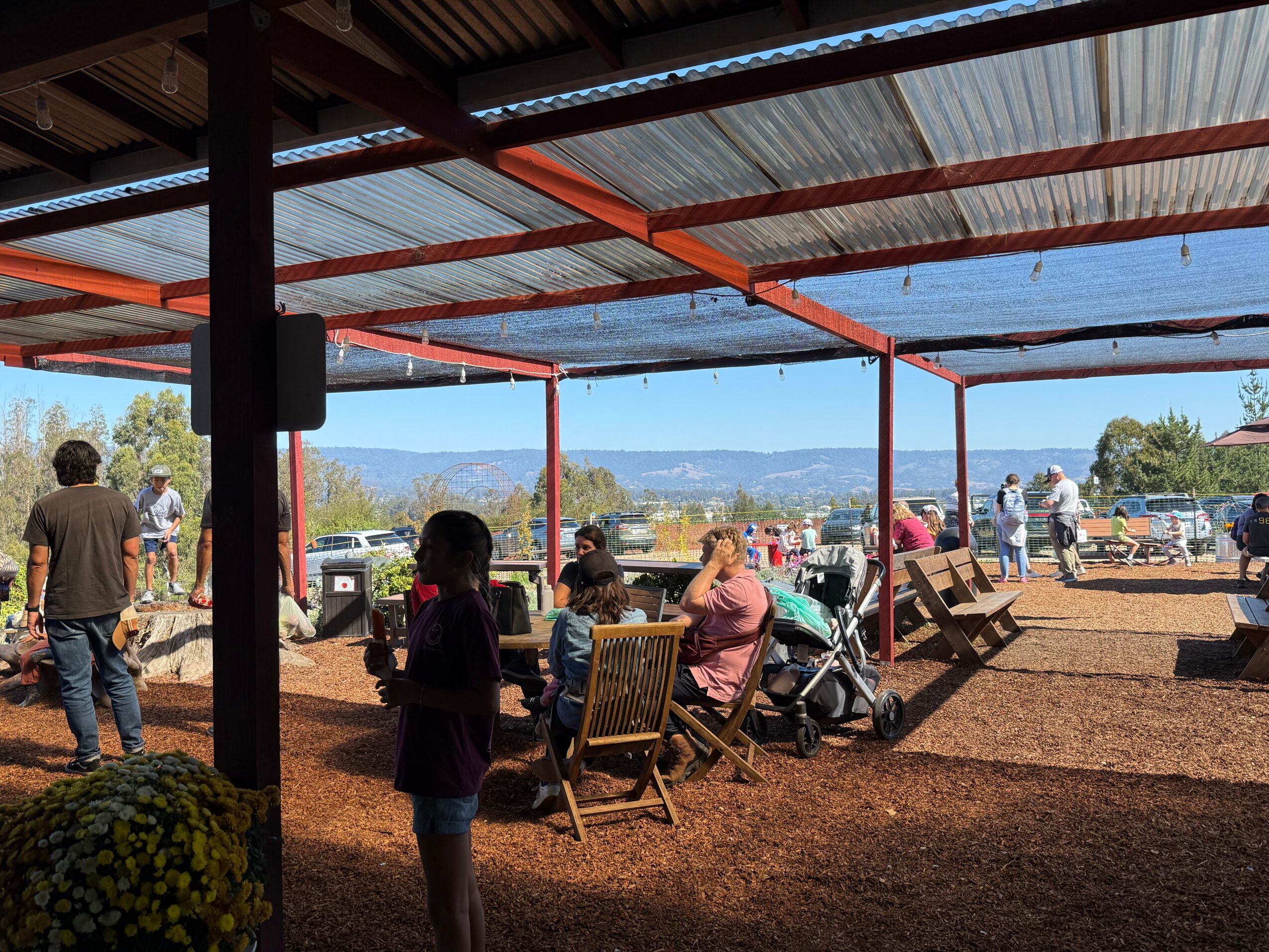 People gather under a metal roof structure outdoors, sitting on chairs and standing, with a scenic landscape in the background.